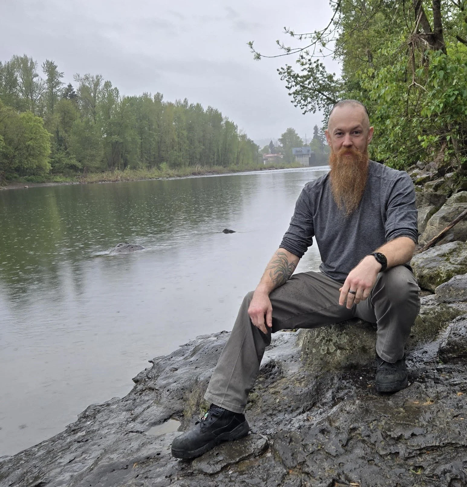 A man with a long beard and tattoos sitting on rocks by a river, surrounded by green trees and overcast sky.