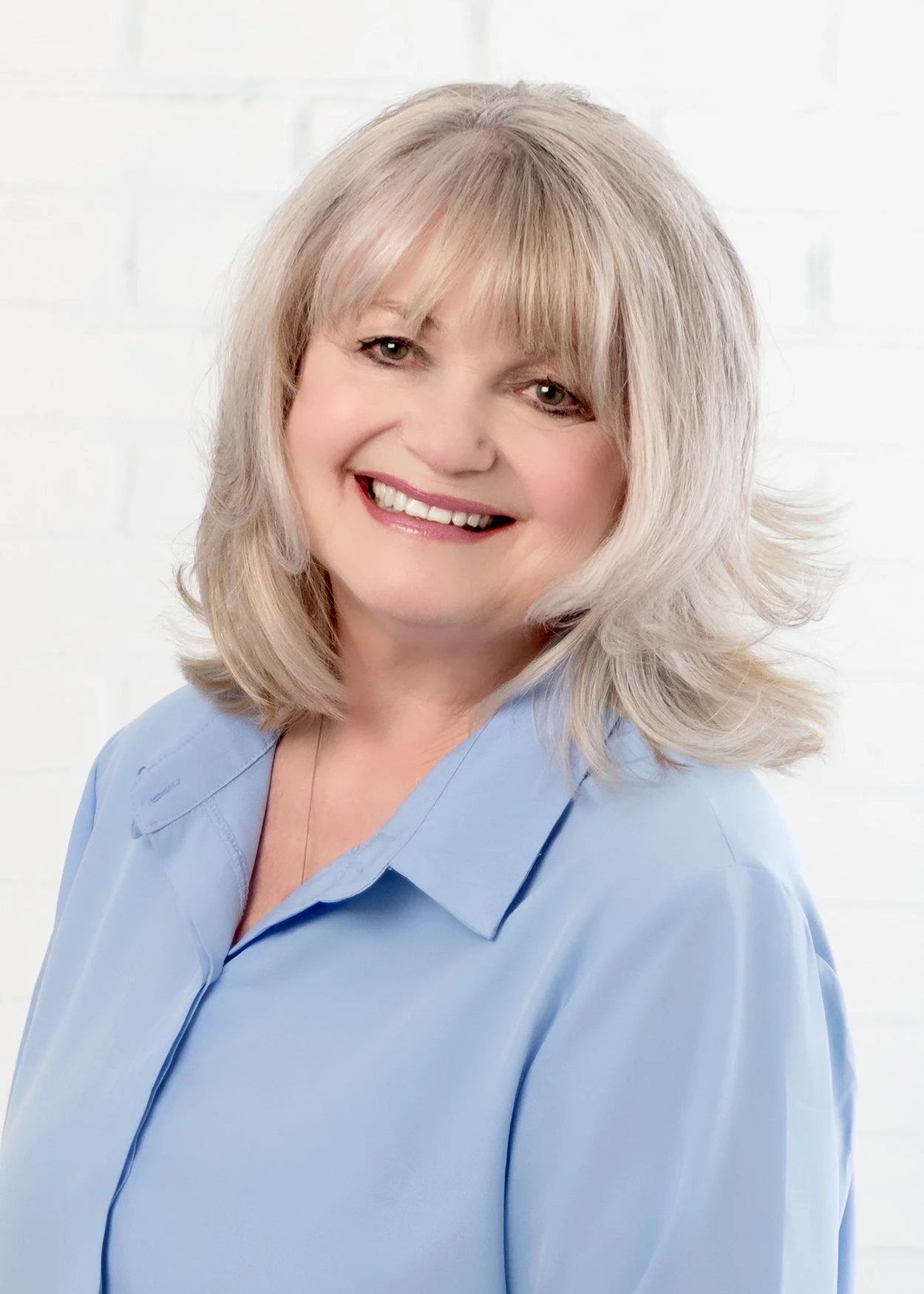 A smiling woman with shoulder-length blonde hair, wearing a light blue shirt, standing against a white background.