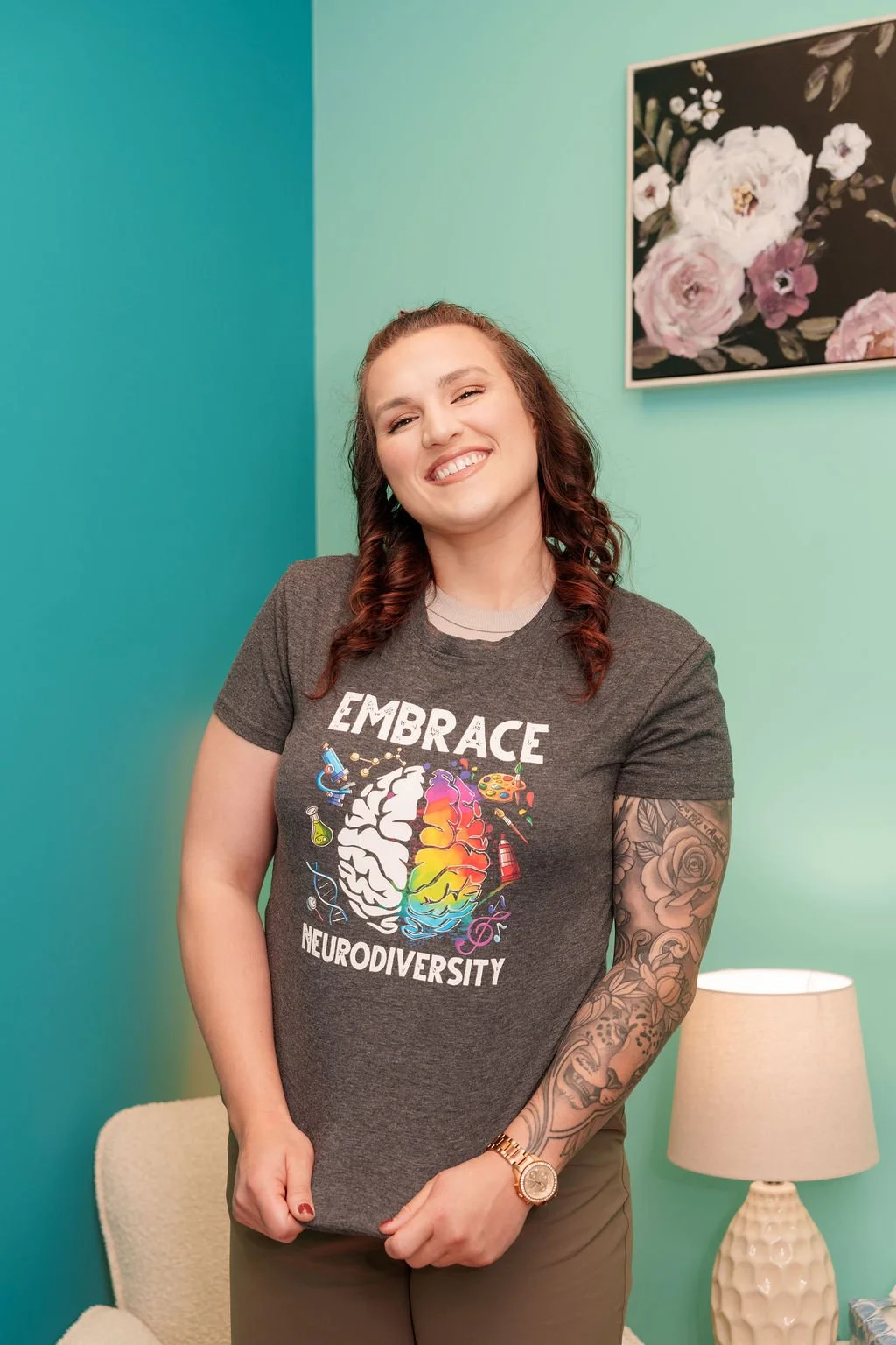 Smiling woman with curly brown hair, tattooed arm, wearing a gray t-shirt with colorful brain and science-themed graphics, standing in a room with teal walls, a floral painting, a beige chair, and a white lamp.