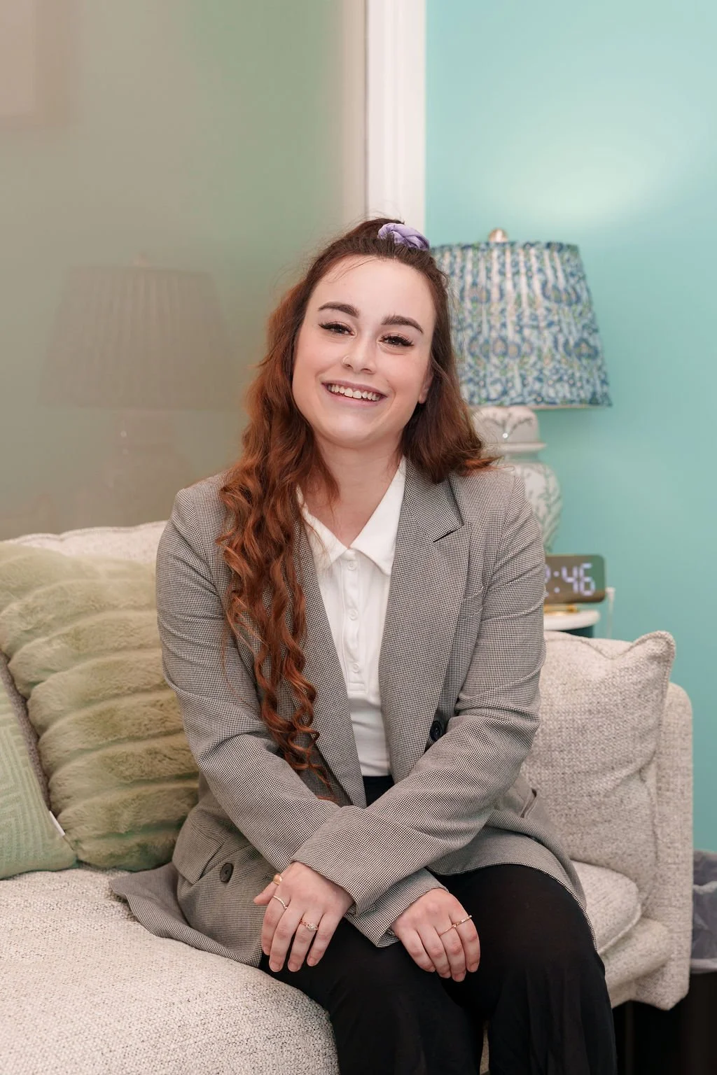 A young woman with long, curly red hair, wearing a gray blazer and white shirt, is sitting on a beige couch with decorative pillows in a cozy living room, smiling at the camera.