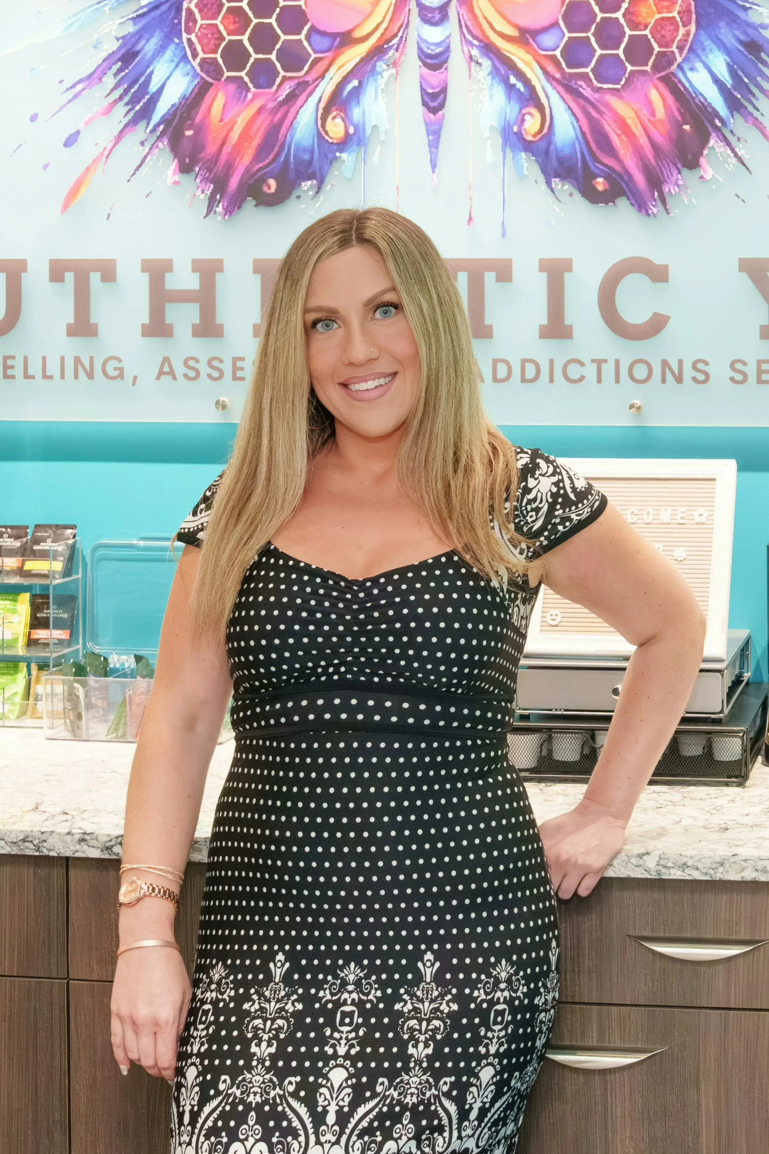 A woman with blonde hair, blue eyes, and light skin stands in front of a colorful background with a butterfly and the word 'AUTHENTIC' displayed prominently. She is wearing a black dress with white polka dots and intricate white patterns near the hem, and she has jewelry on her left wrist and a nose piercing. The background features a counter with various items and a wall with text about healing and addiction services.