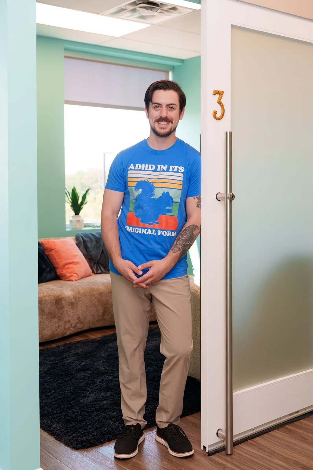 A young man standing in a doorway inside an office or therapy room, smiling at the camera. He has dark hair, a beard, and tattoos on his left arm. He is wearing a bright blue T-shirt with an illustration and the text 'ADHD IN ITS ORIGINAL FORM,' beige pants, and black shoes. Behind him, there is a room with a couch and a window with greenery outside.