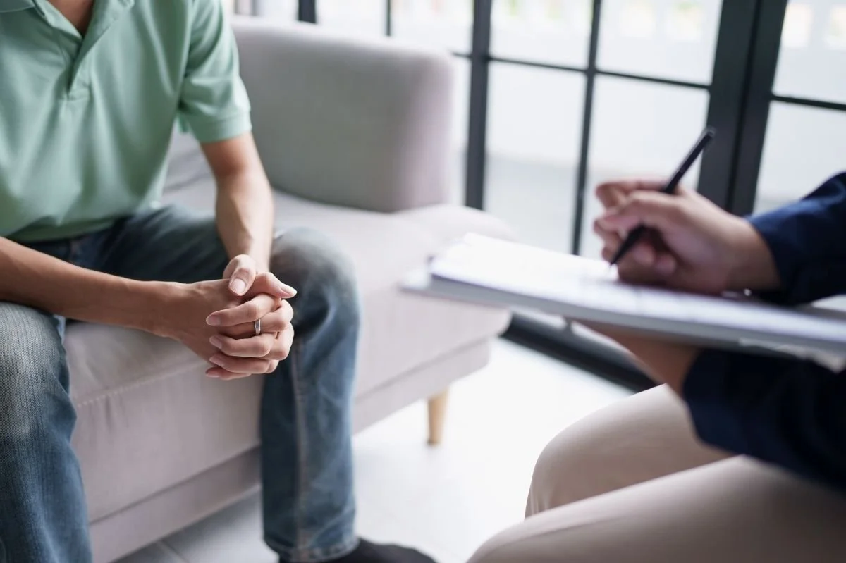 Male client sitting on a couch with hands clasped while therapist writes notes during therapy session.