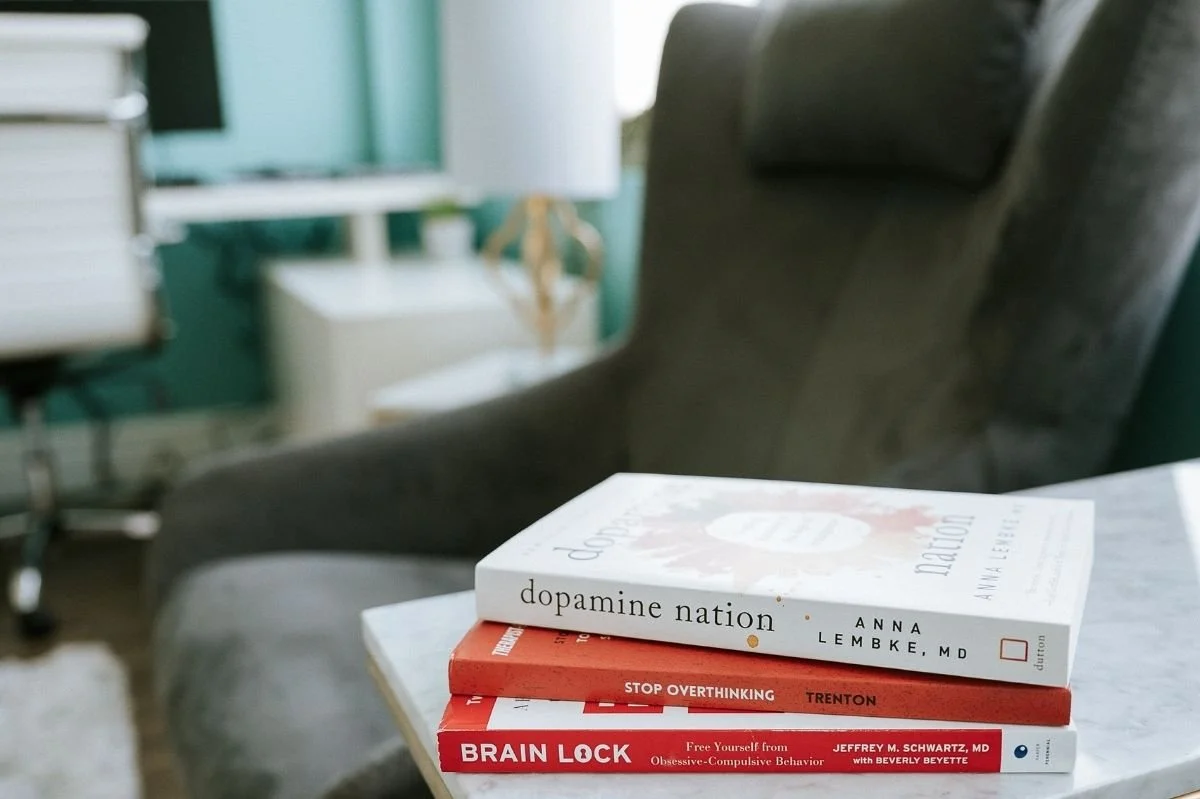 Stack of psychology books including Dopamine Nation and Brain Lock on a table in a therapy office.