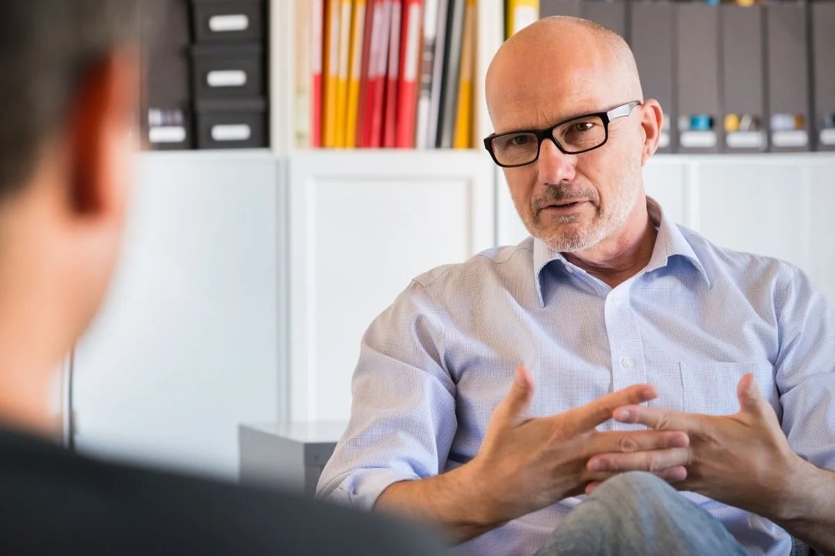 Male therapist gesturing with hands while explaining something to client in office.