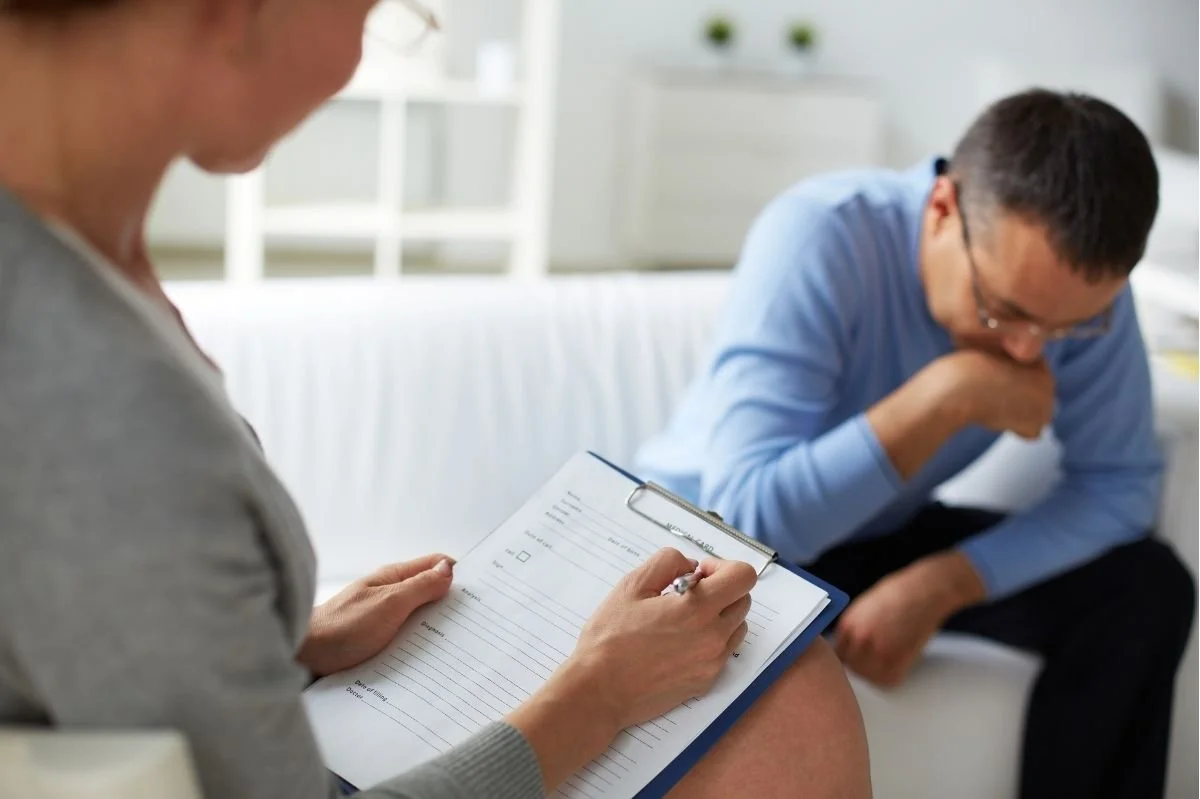 Therapist writing notes on clipboard while male client sits with head lowered during counseling session.
