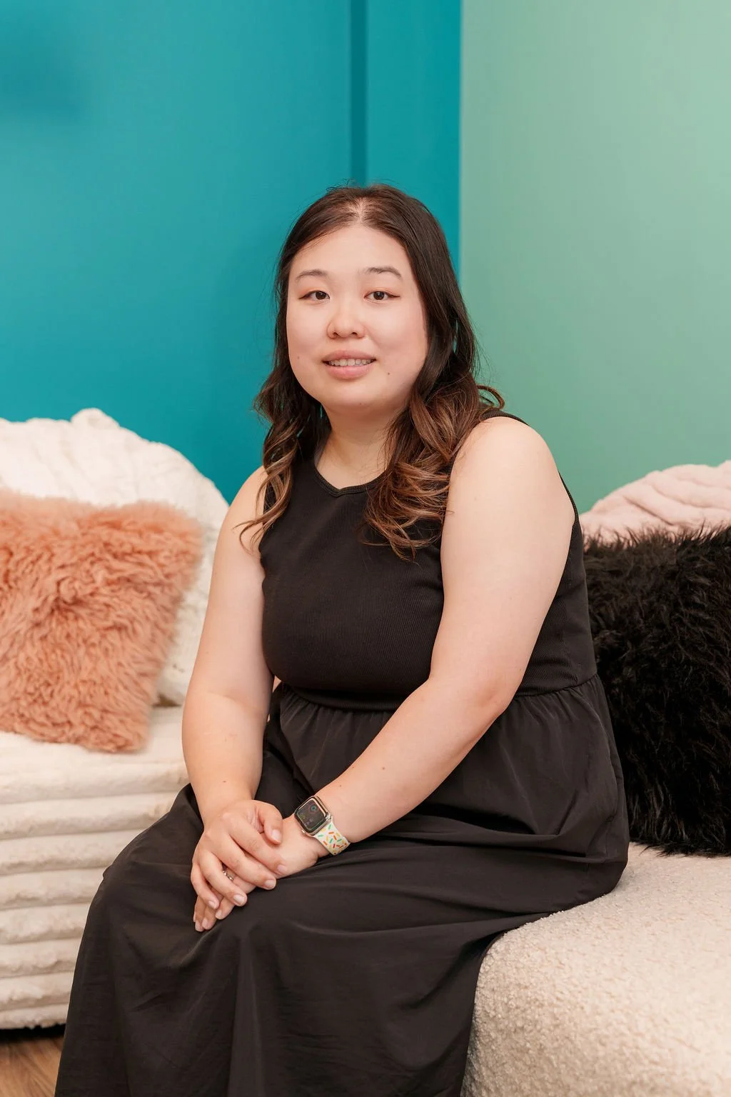 A woman with long brown hair, sitting on a beige couch with a black dress, smiling at the camera, with a teal wall and fluffy pillows in the background.