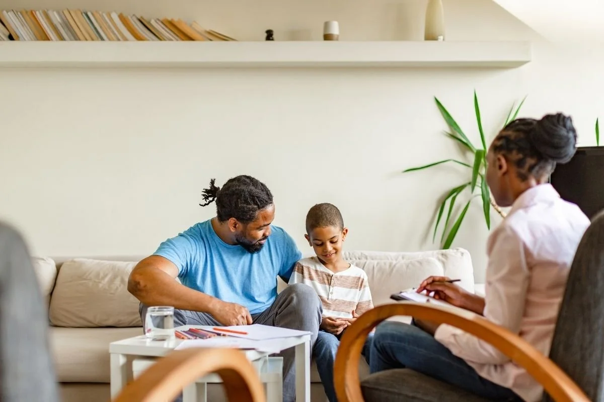 Father and young son participating in family therapy session with counselor in a comfortable office setting.