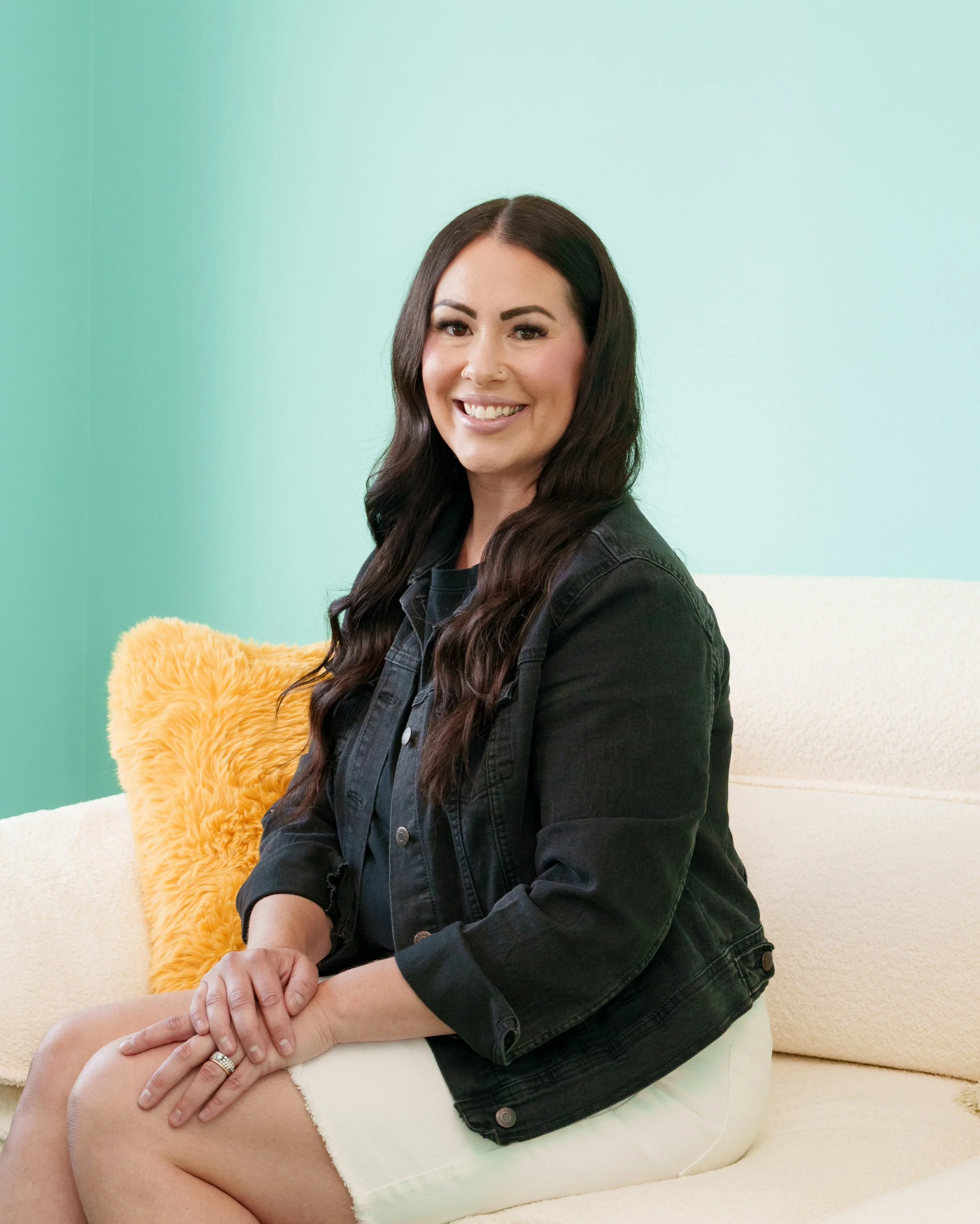 A woman with long dark hair smiling, sitting next to a person lying on a cream-colored sofa, with a green wall in the background and a fluffy yellow pillow nearby.