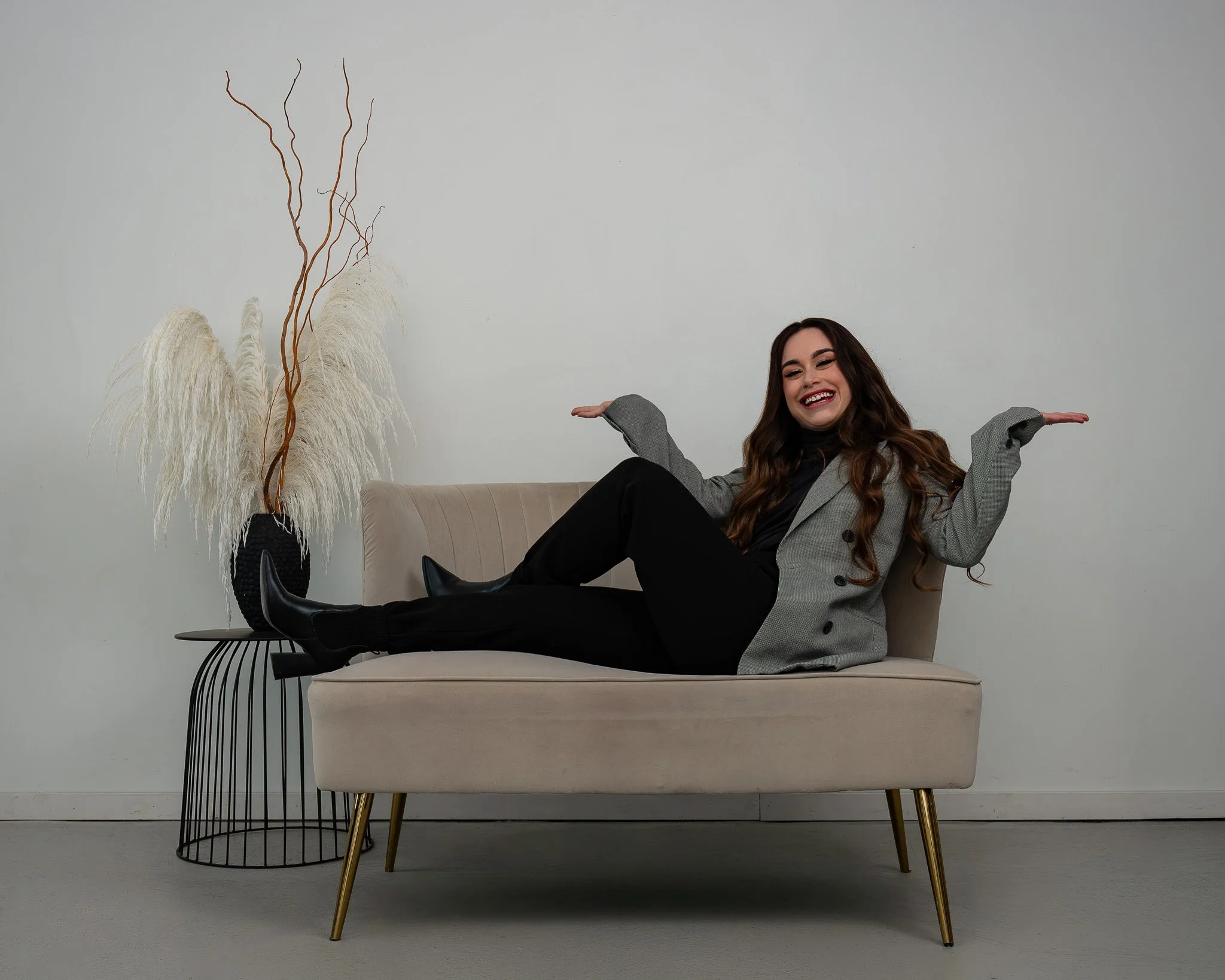 A woman with long brown hair and a broad smile sitting on a beige sofa with gold legs, wearing a gray blazer and black pants, raising her arms with palms facing upward. To her left, there is a black wire side table with a large black vase filled with tall, wavy dried branches and white pampas grass, set against a plain light gray wall.