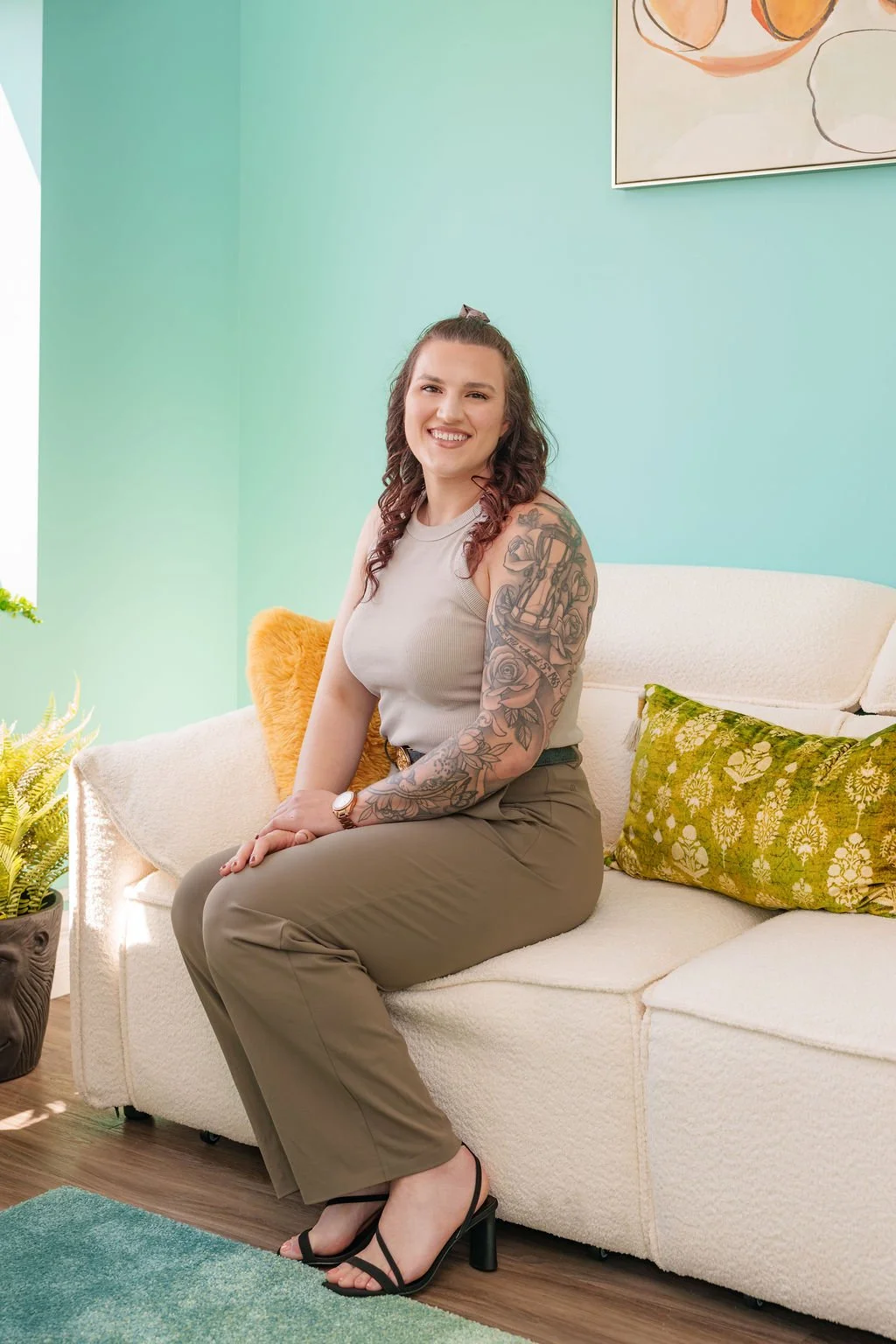 A woman with curly brown hair, smiling, sitting on a white sofa in a brightly lit living room, wearing a sleeveless beige top, khaki pants, and black high heels, with a visible tattoo sleeve on her left arm.