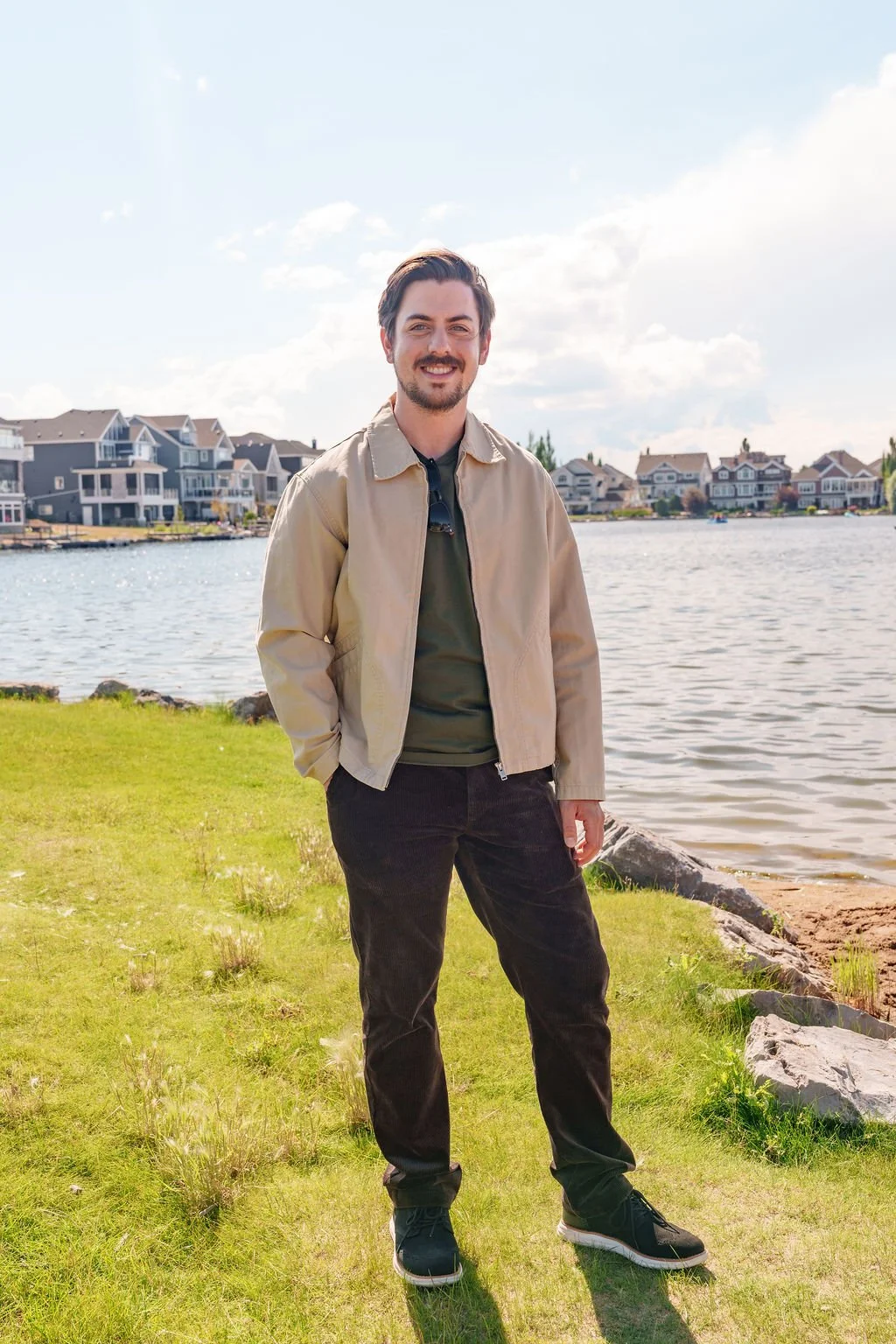 A smiling man standing by a lakeside on a sunny day with houses in the background.