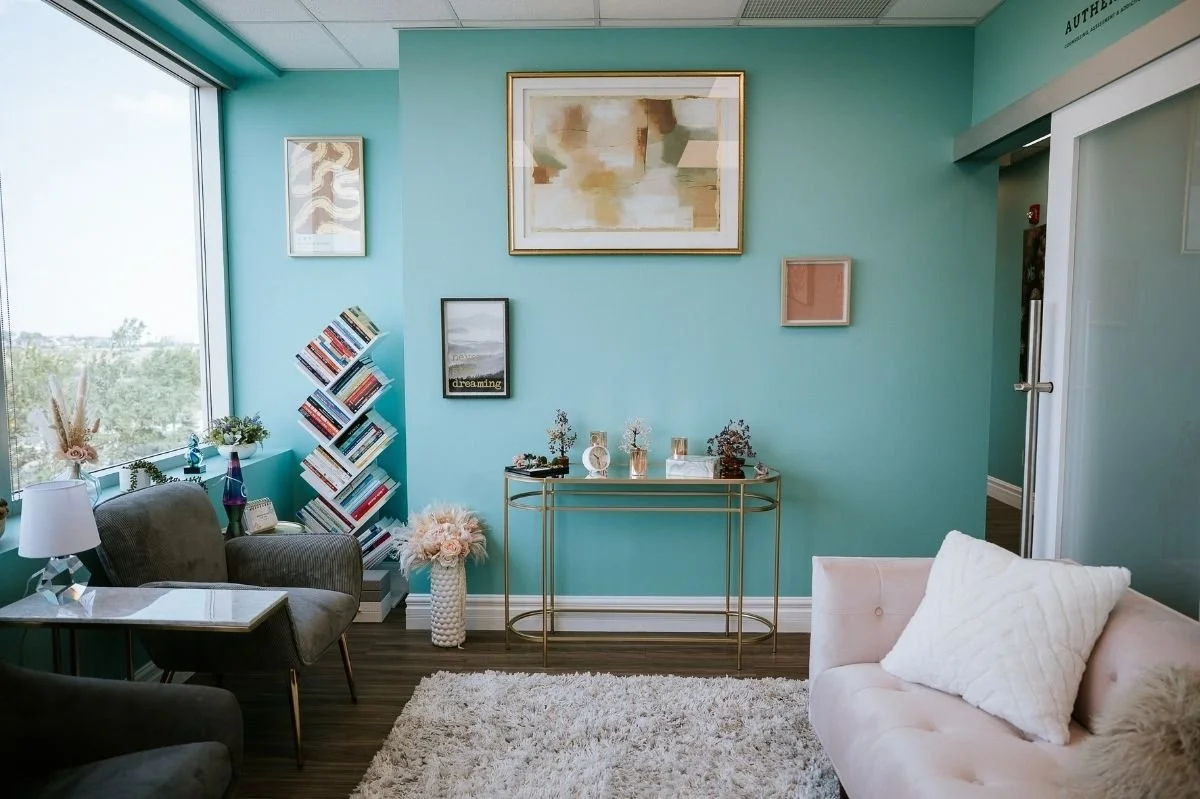 Modern therapy office waiting room with teal walls, abstract art, bookshelf, and soft seating.