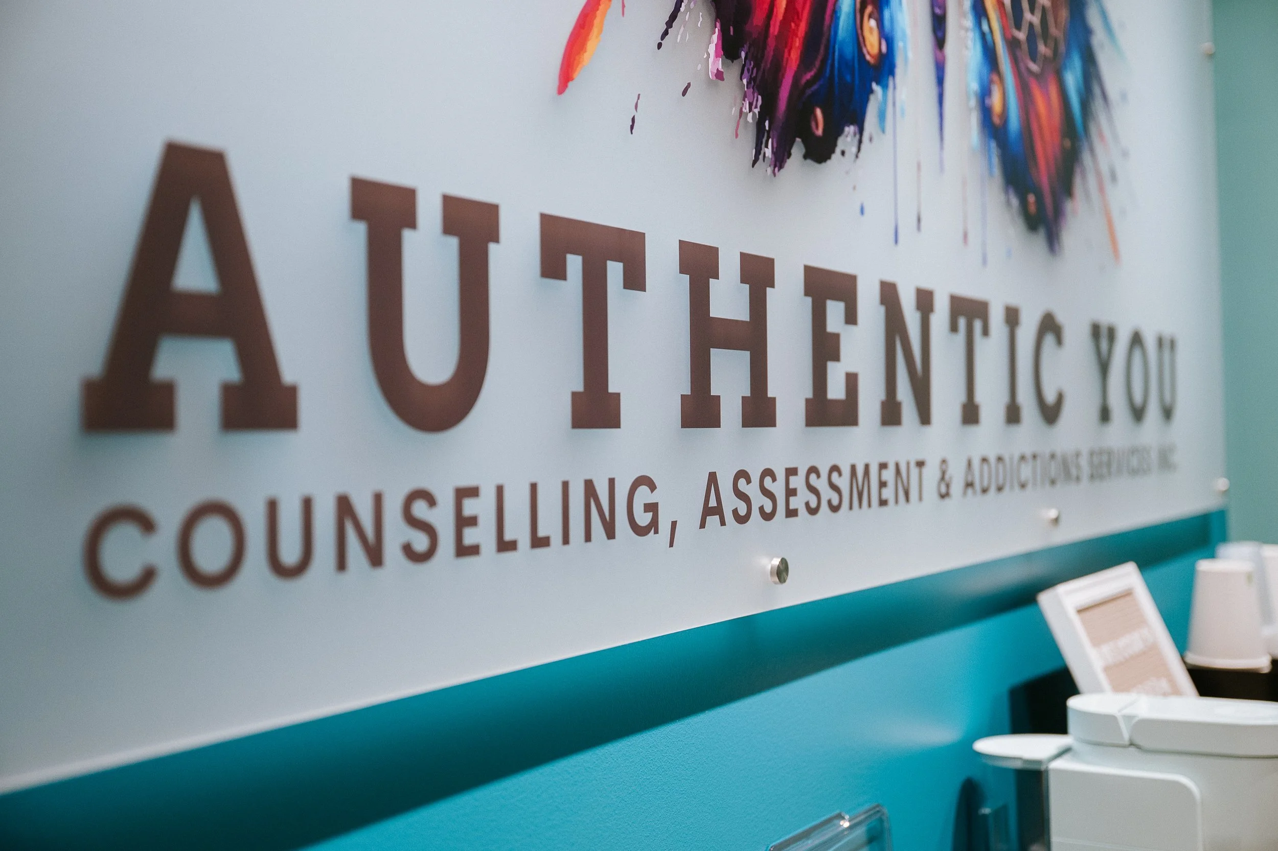 Wall sign reading Authentic You Counselling, Assessment and Addictions Services inside therapy clinic reception area.