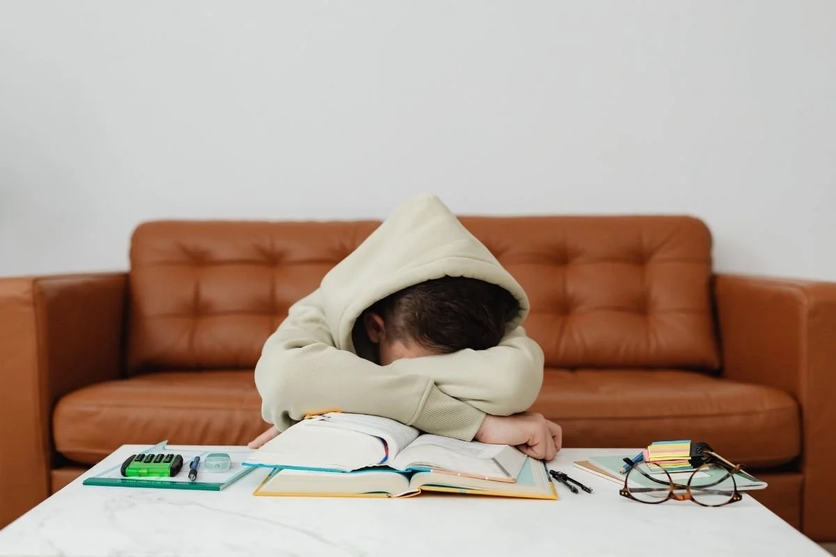 Teen student in hoodie resting head on arms over open books, appearing overwhelmed while studying at home.