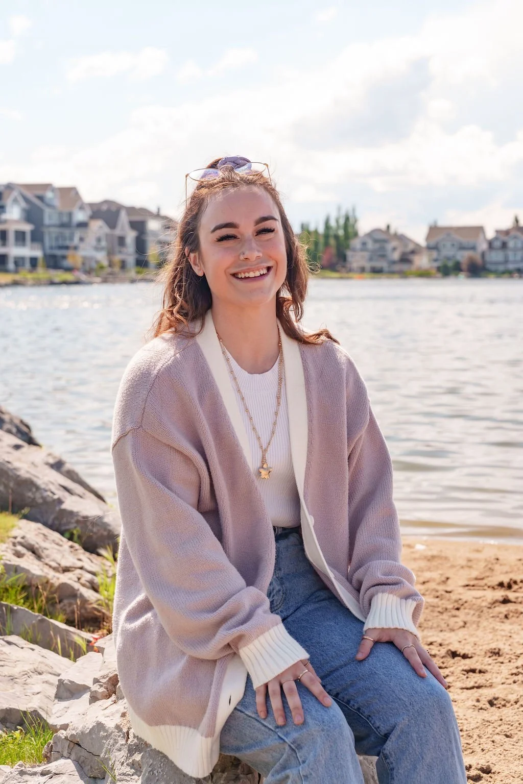 Young woman sitting on rocks by a lake, smiling, with houses and trees in the background.