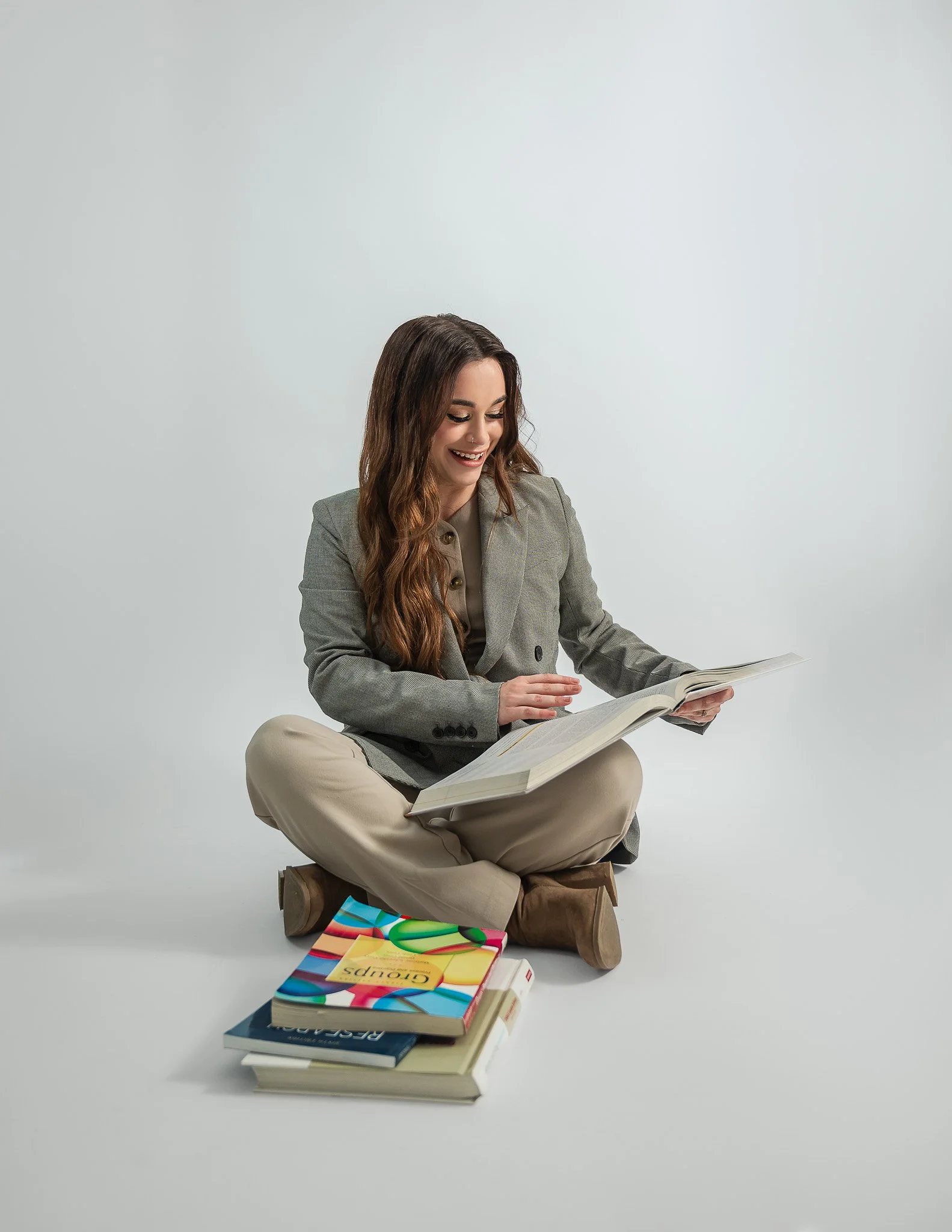 A woman sitting cross-legged on a small wooden stool, reading a book. She has long, wavy brown hair and is wearing a gray blazer and beige pants. Multiple large books are stacked on the floor in front of her, and colorful magazines are on top of the stack. The background is plain and white.