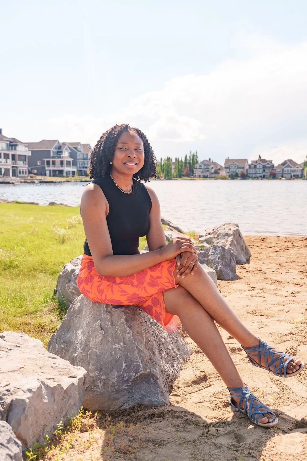 A woman sitting on a rock by a lake, with residential houses in the background, wearing a black sleeveless top, orange skirt, and blue sandals, smiling at the camera.