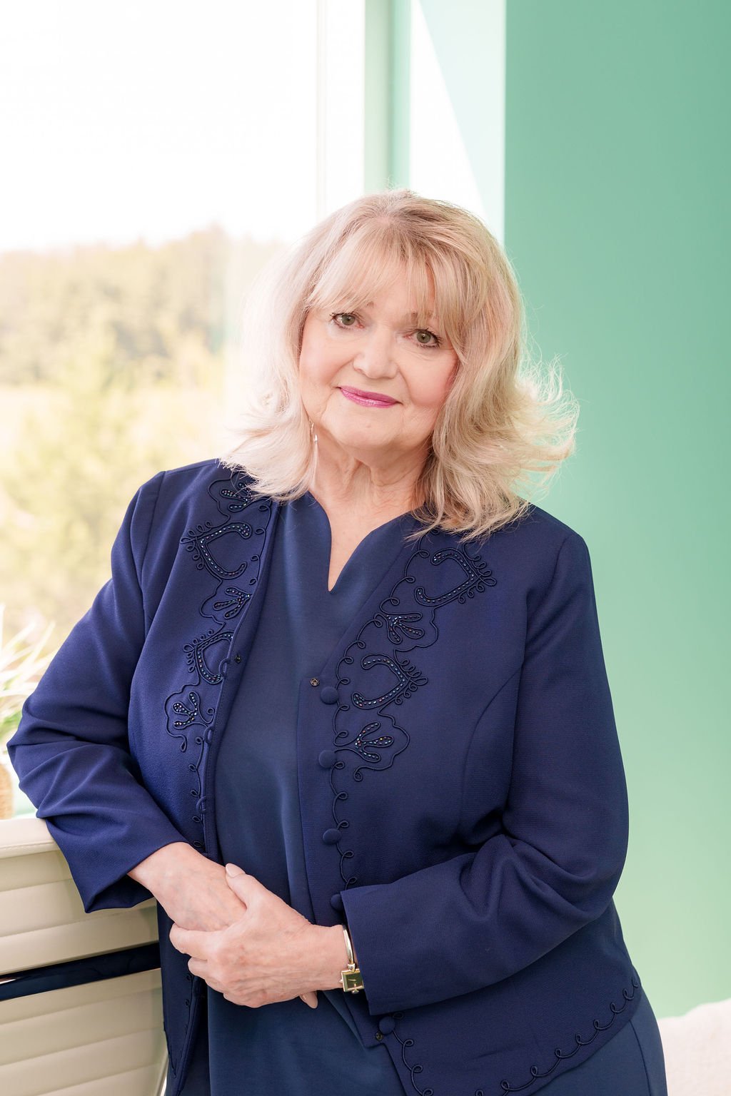 A woman with shoulder-length blonde hair, wearing a dark blue blazer with embroidery, standing indoors near a window with a green wall background.