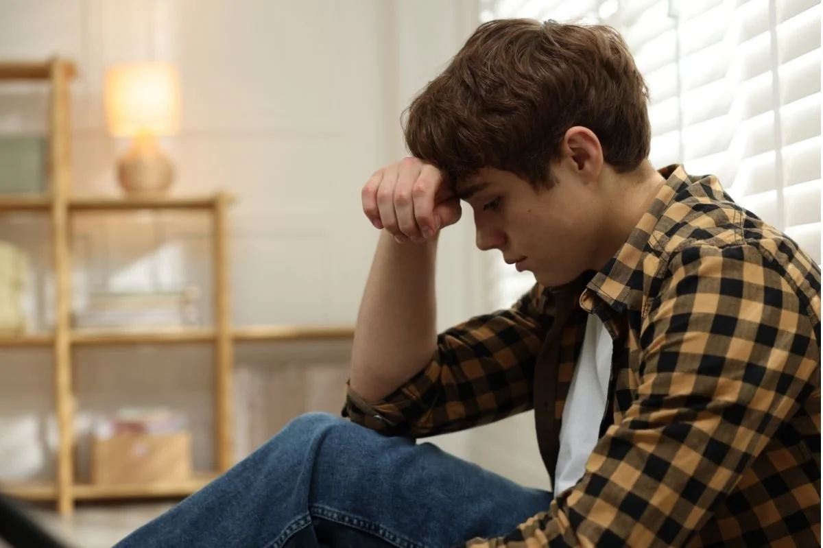 Teenage boy sitting alone with head resting on hand, appearing worried or anxious in a home setting.