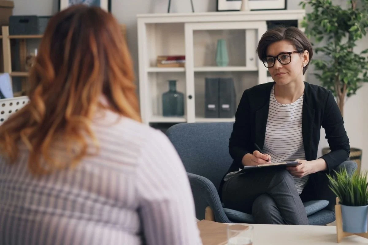 Therapist holding a clipboard and listening attentively to a client seated on a couch.