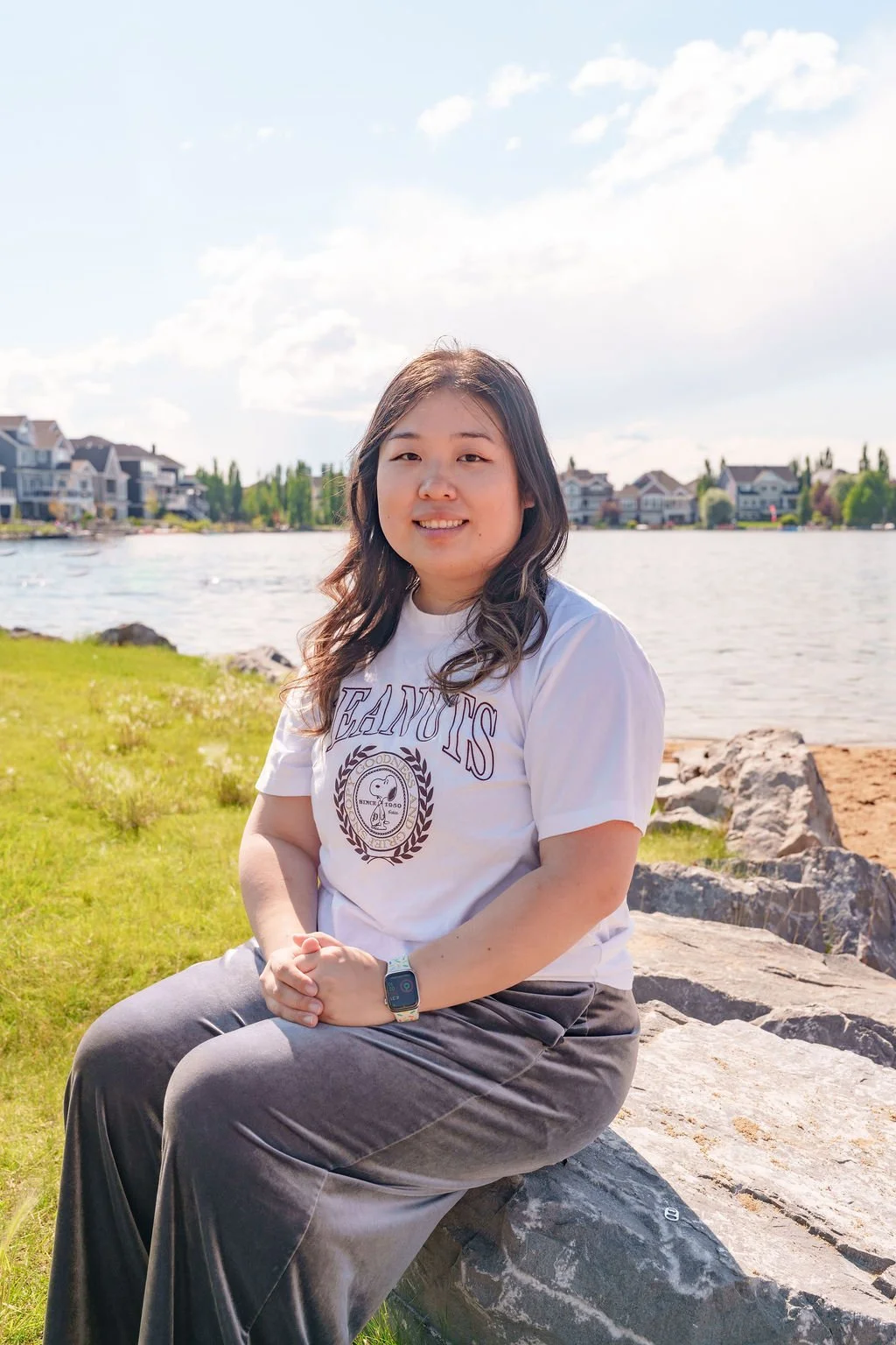 A woman sitting on rocks near a lake with suburban houses in the background, under a partly cloudy sky.
