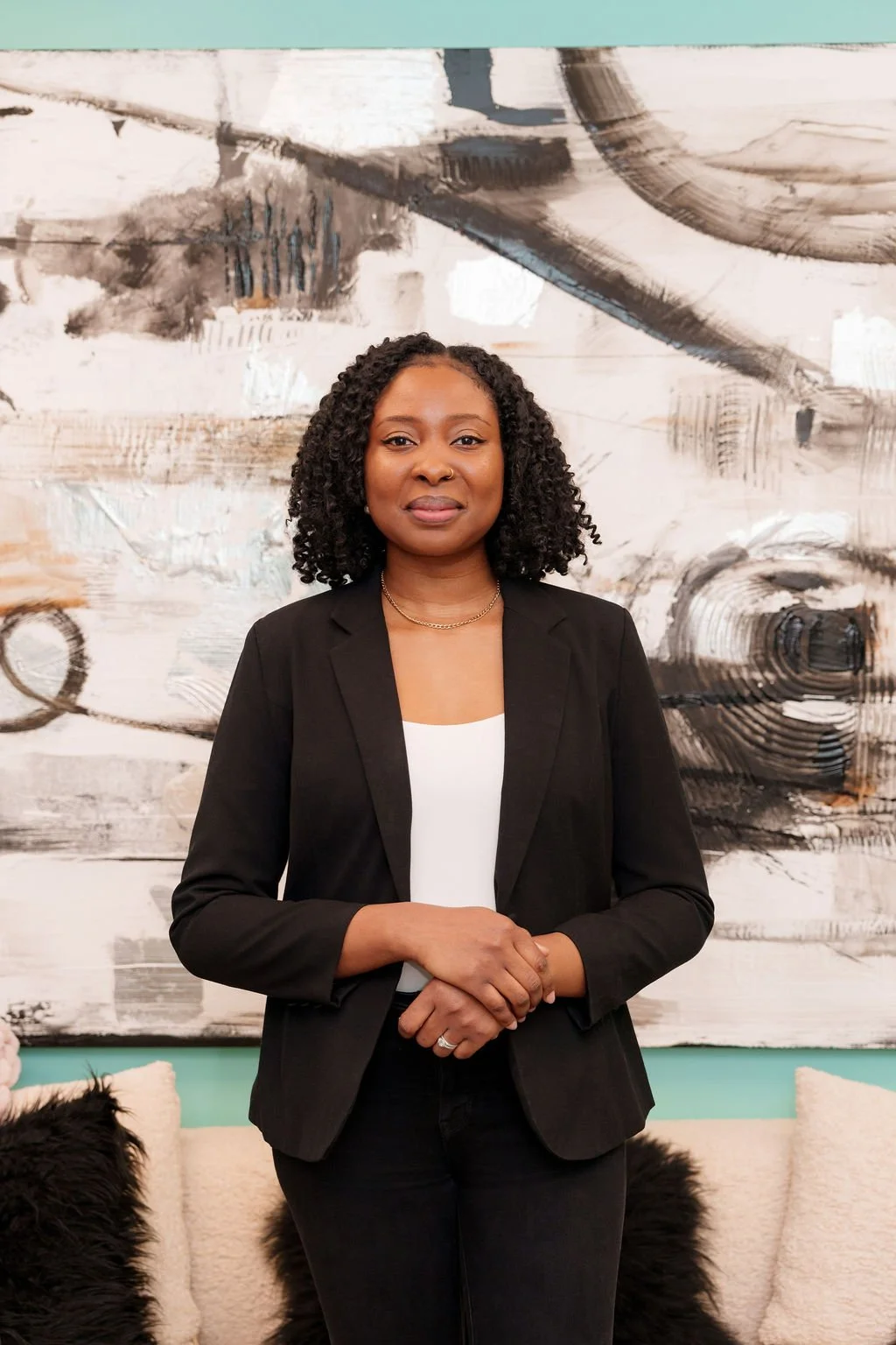 A woman with curly black hair, wearing a black blazer over a white top, standing indoors in front of an abstract painting.