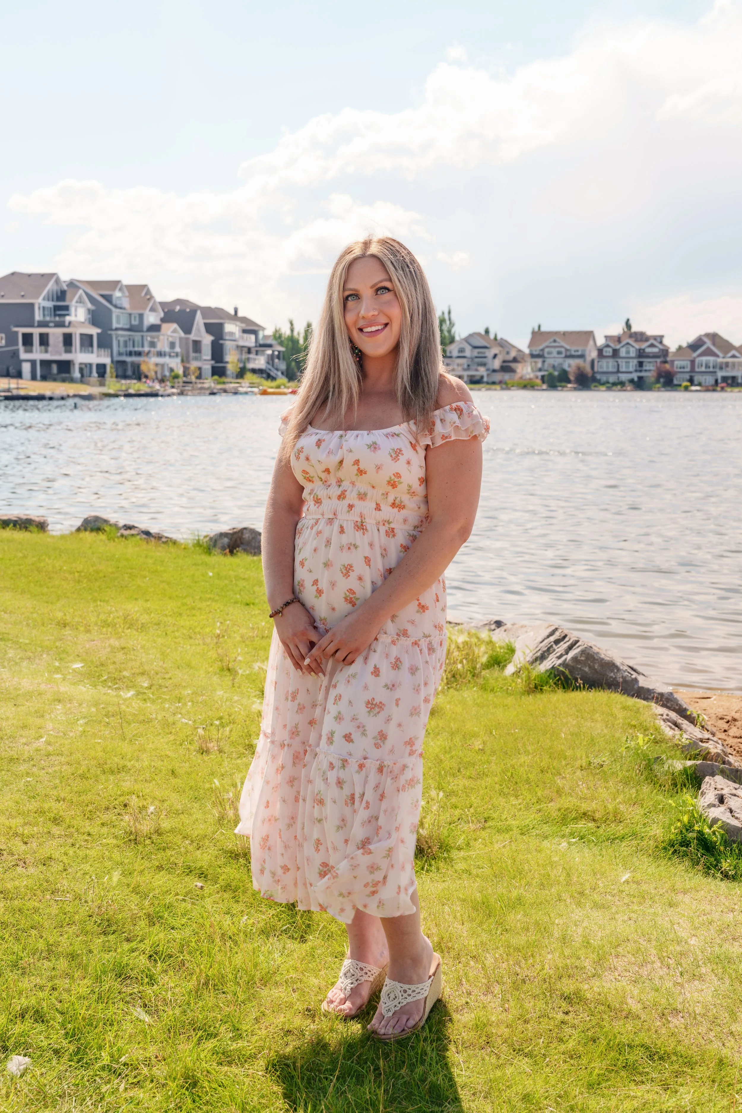 A woman with blonde hair wearing a long floral dress and lace sandals standing on grass next to water with houses in the background.