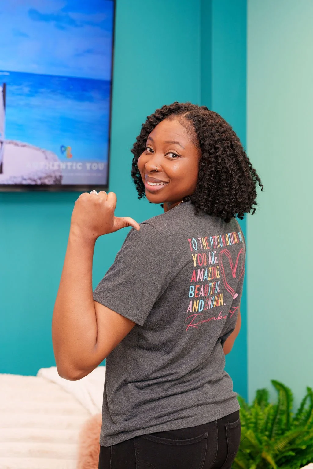 A smiling woman with curly hair pointing to her shoulder, wearing a gray T-shirt with colorful text on the back, standing in a room with teal walls and a TV screen.
