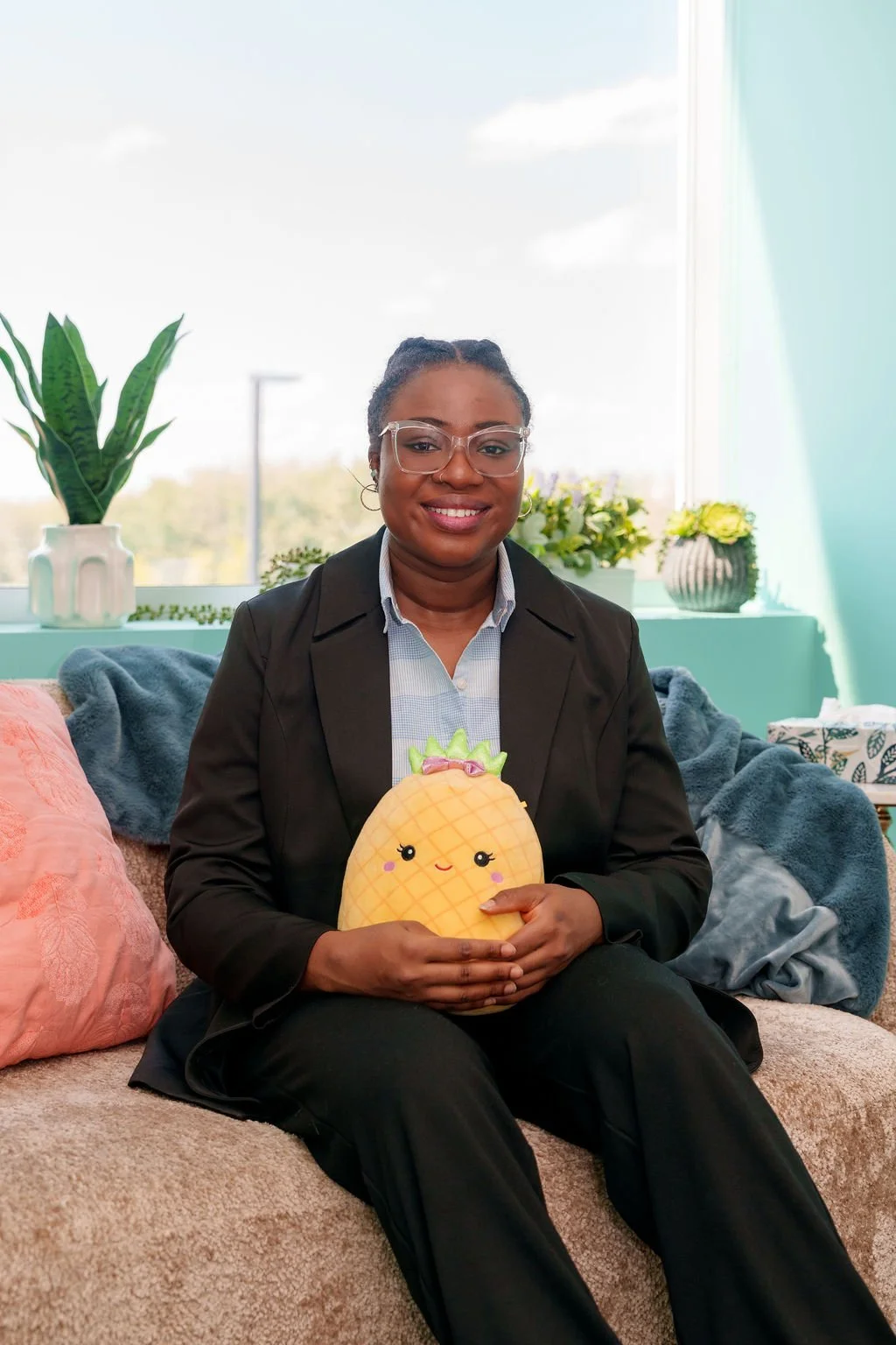 A woman in glasses and business attire sitting on a couch, holding a plush pineapple pillow with a cute face, in a bright room with plants and a large window.