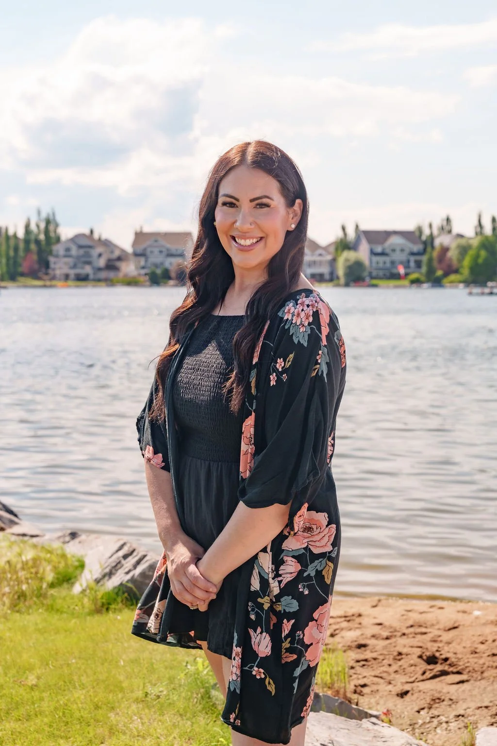 A woman with long dark hair smiling at the camera while standing near a body of water, with houses visible in the background.