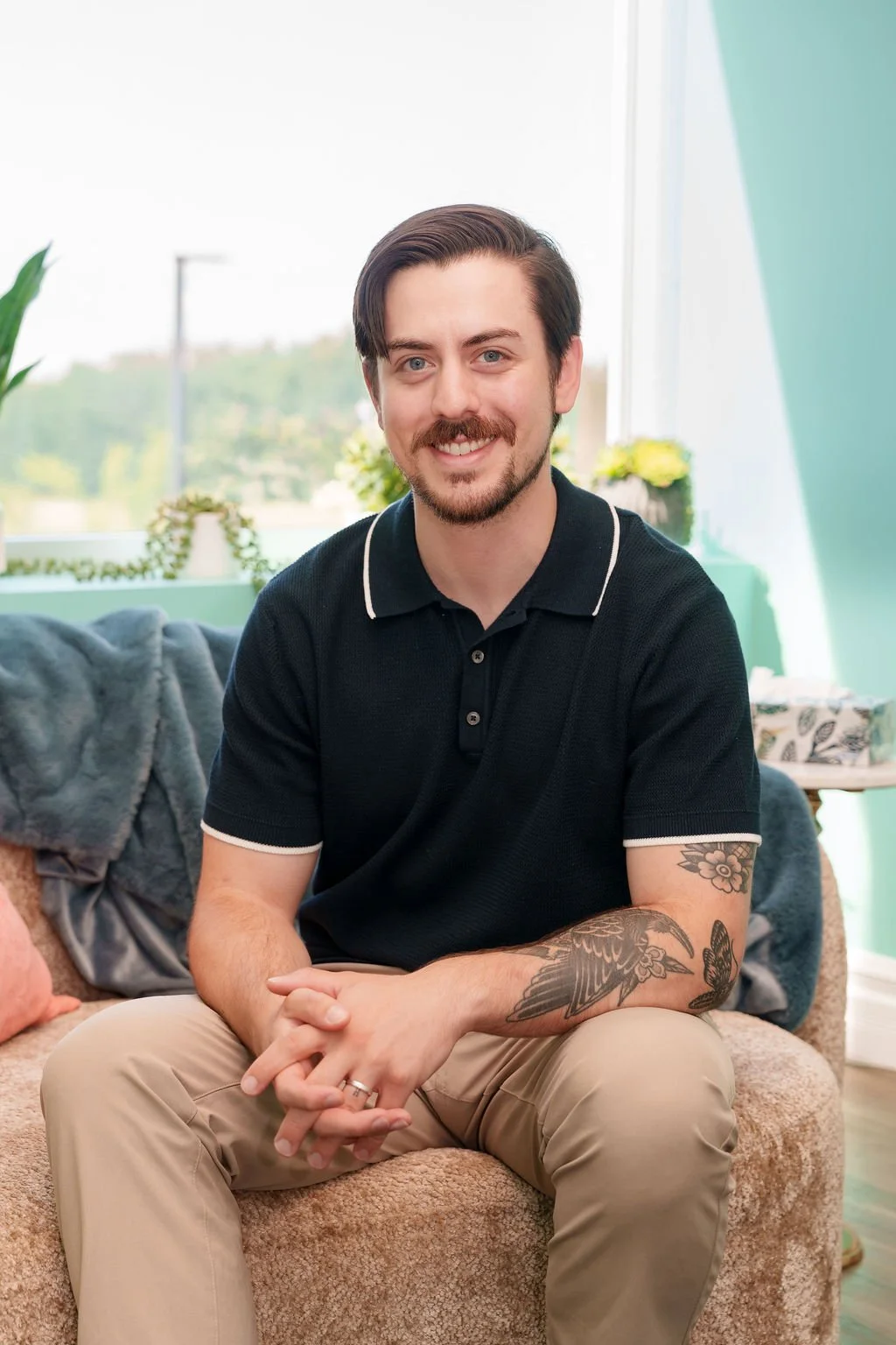 A young man with dark hair, a beard, and tattoos, sitting on a beige couch in a well-lit room with large windows and green walls, smiling at the camera.