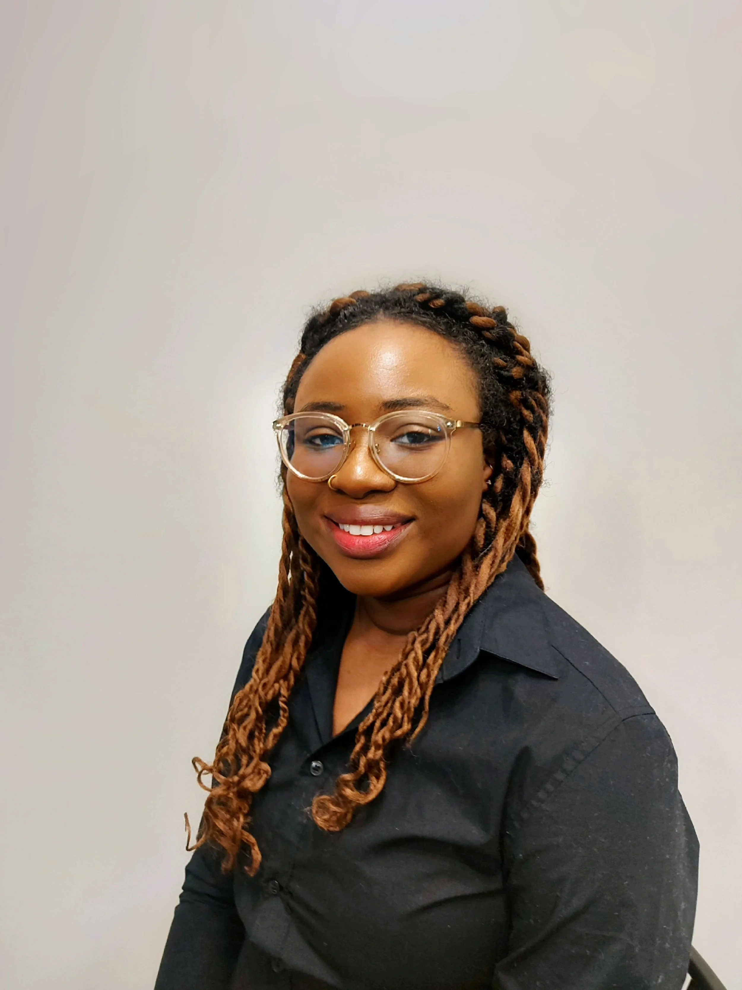 A woman with braids and glasses smiling, wearing a black shirt, standing against a plain white wall.