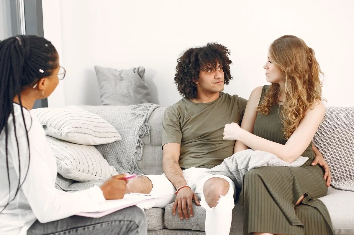 Couple sitting close together on couch while therapist takes notes during counseling session.
