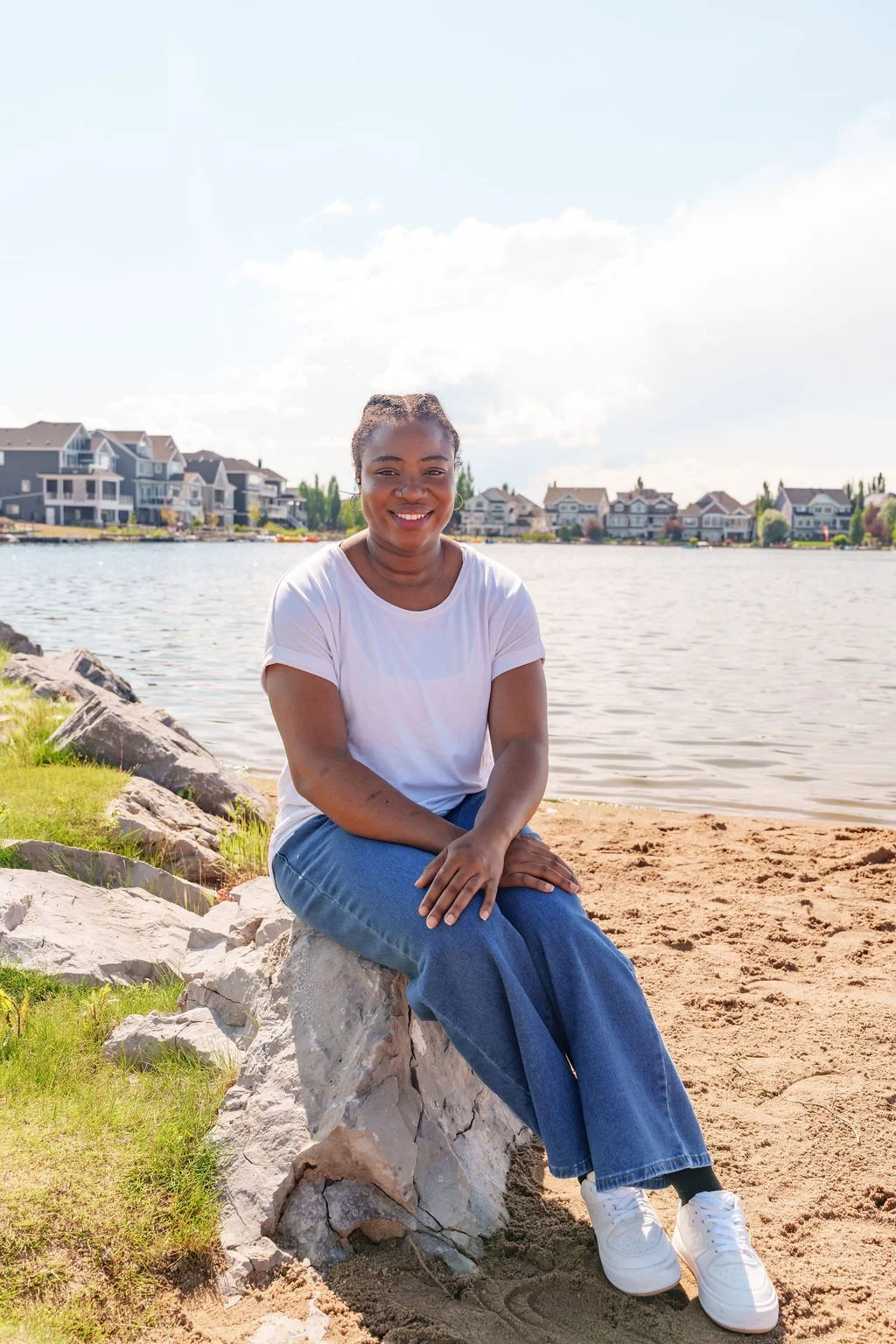 A woman sitting on rocks by a lake with houses in the background, smiling at the camera.