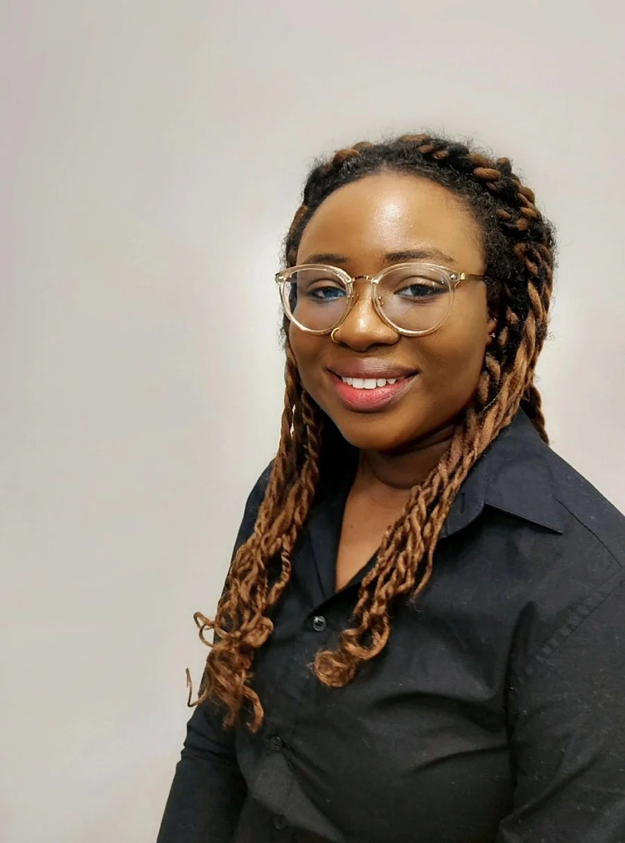 Portrait of a smiling woman with glasses and braided hair, wearing a black shirt, against a plain background.
