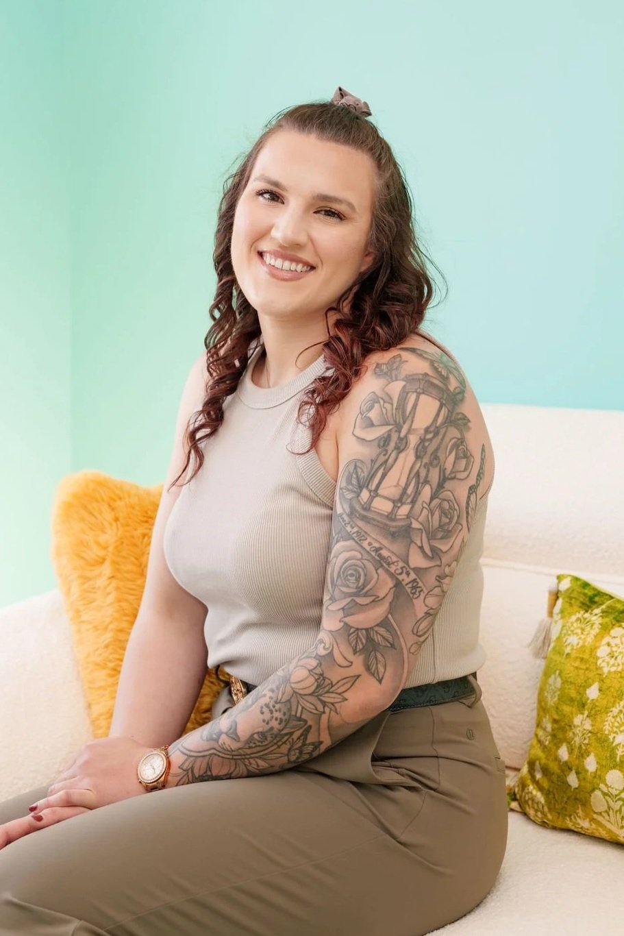 A woman with long curly hair, wearing a gray sleeveless top and khaki pants, sitting on a white couch with green and yellow pillows, smiling at the camera in a brightly lit room with pastel green walls.