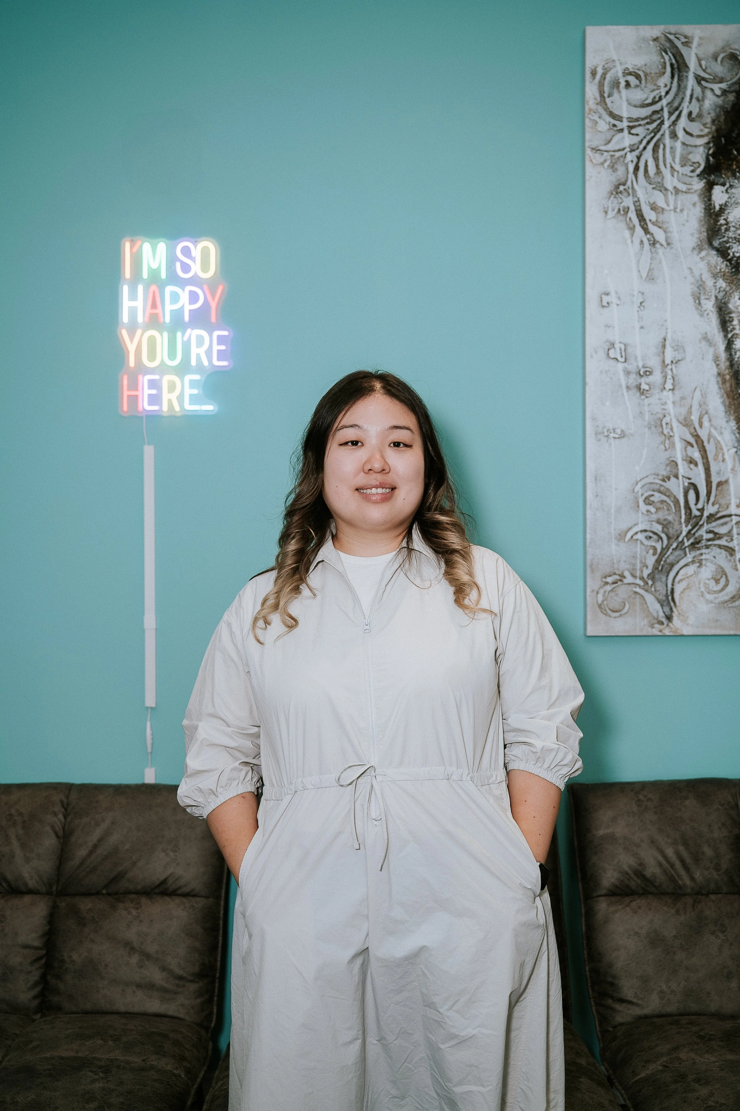 A woman with dark hair and light highlights smiling, wearing a white zip-up jacket with a drawstring waist, standing in front of a teal wall with a colorful neon sign that reads 'I'M SO HAPPY YOU'RE HERE,' and a large black and white artistic painting on the right side, with two dark brown sofas at the bottom of the image.