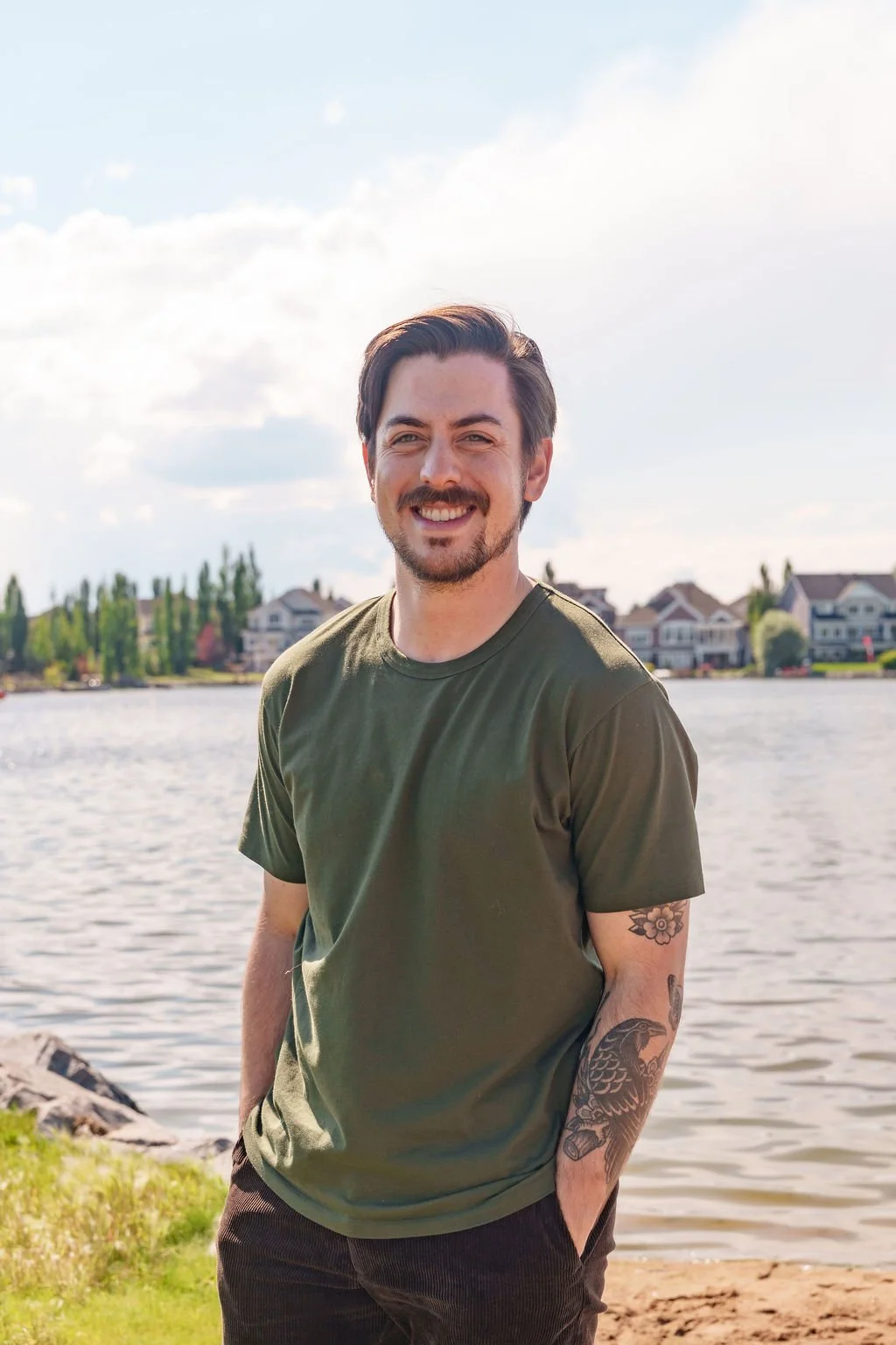 A smiling man with dark hair and a beard standing near a body of water on a sunny day, with houses and trees in the background.