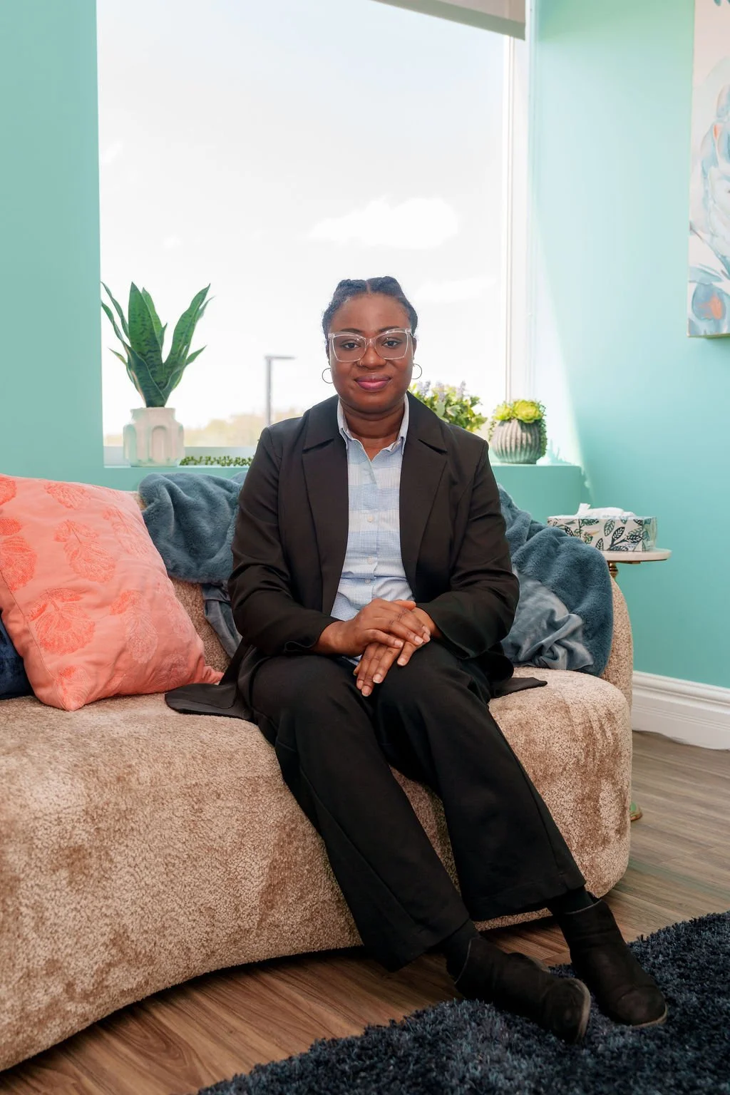 A woman sitting on a beige sofa with pink and blue pillows in a room with aqua-colored walls. She is wearing glasses, a black blazer, a light blue shirt, black pants, and black shoes. A window behind her shows a bright sky with some clouds, and there are potted plants and flowers on the windowsill.