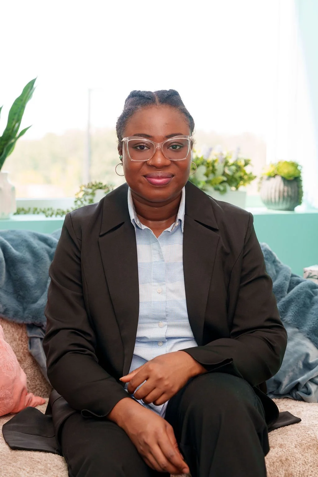 A woman sitting on a sofa in an indoor setting with plants in the background, wearing glasses, a black blazer, and a light-colored button-up shirt.
