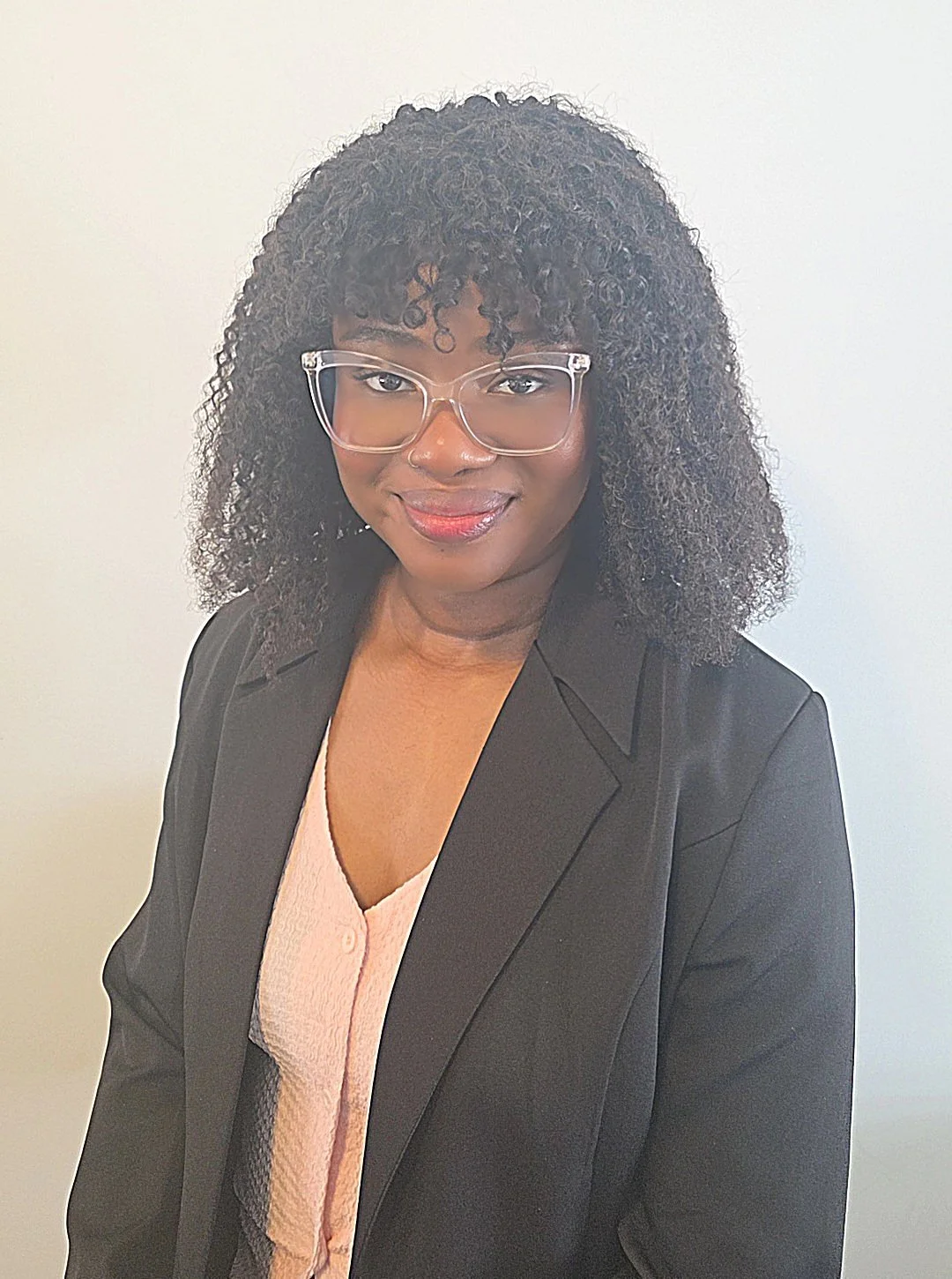 Portrait of a woman with curly hair, wearing glasses, a black blazer, and a light pink top, smiling against a plain white background.