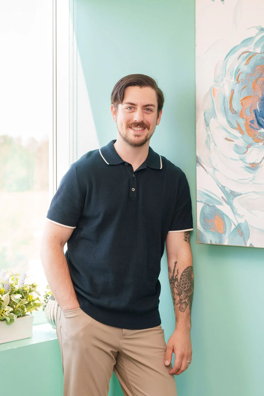 A young man with dark hair and a beard, wearing a navy polo shirt and khaki pants, standing indoors next to a green wall and a colorful abstract painting.