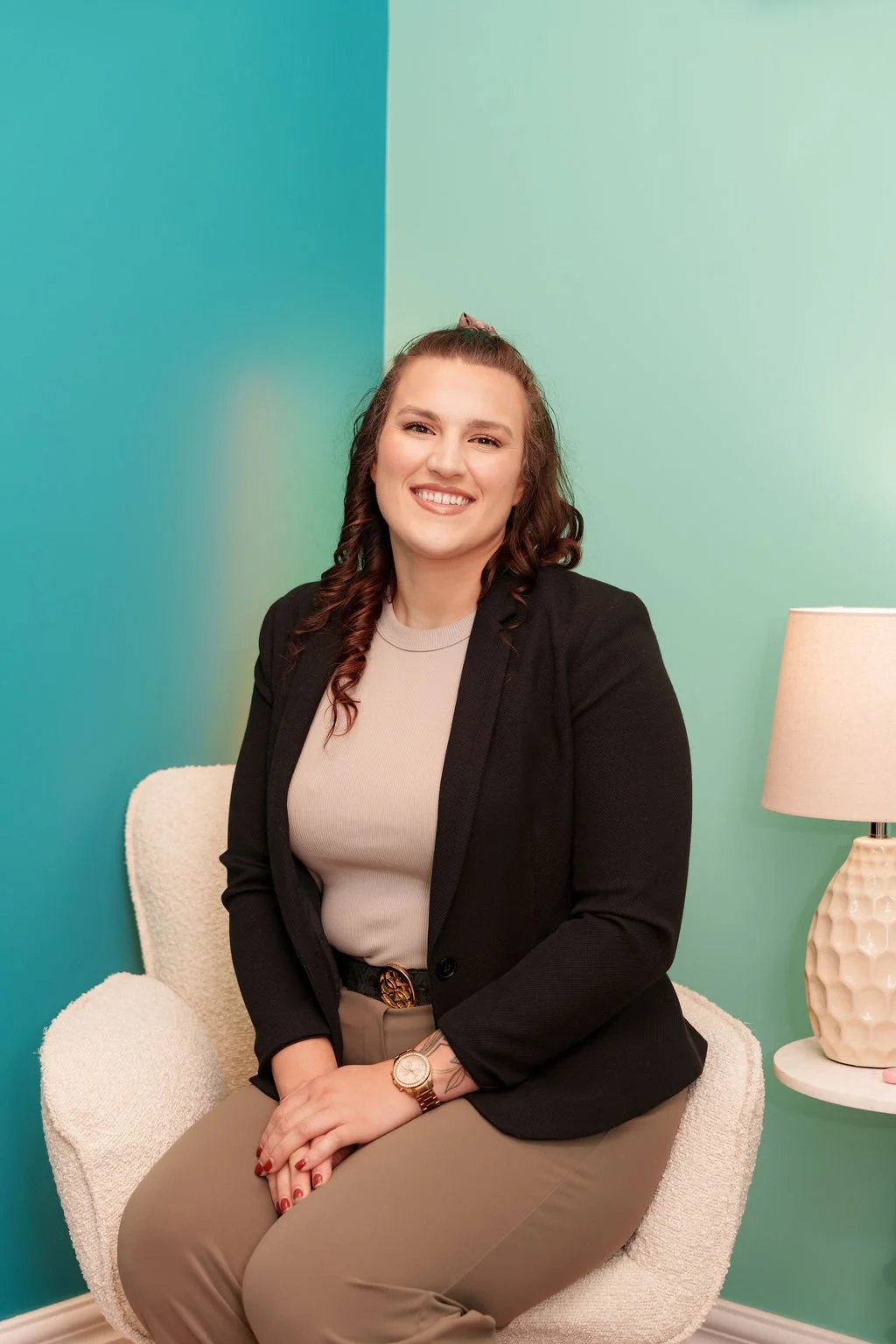 A woman sitting on a cream-colored chair with a textured surface, smiling, wearing a beige top, black blazer, and beige pants. She has brown, curly hair and wears a gold watch and ring. Beside her, there is a white side table with a cream-colored textured lamp, set against a two-tone green and teal wall.