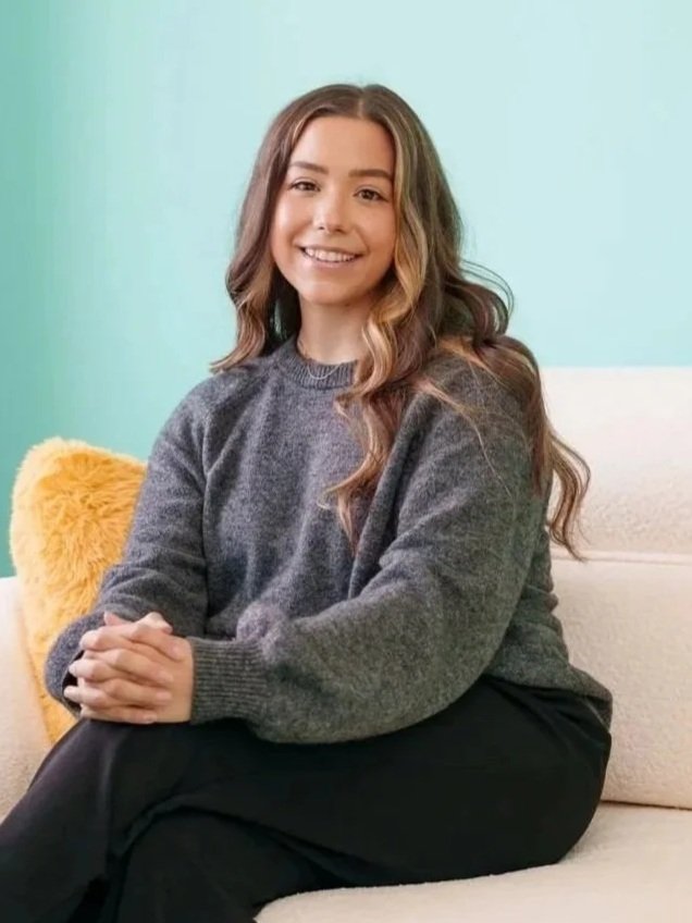 A young woman with long wavy hair smiling while sitting on a cream-colored sofa with a yellow fluffy pillow against a light blue wall.