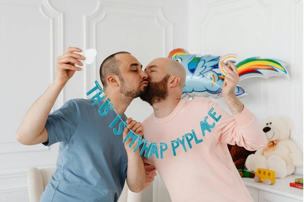 Two men kissing while holding a "This Is My Happy Place" banner in a joyful pregnancy announcement setting.