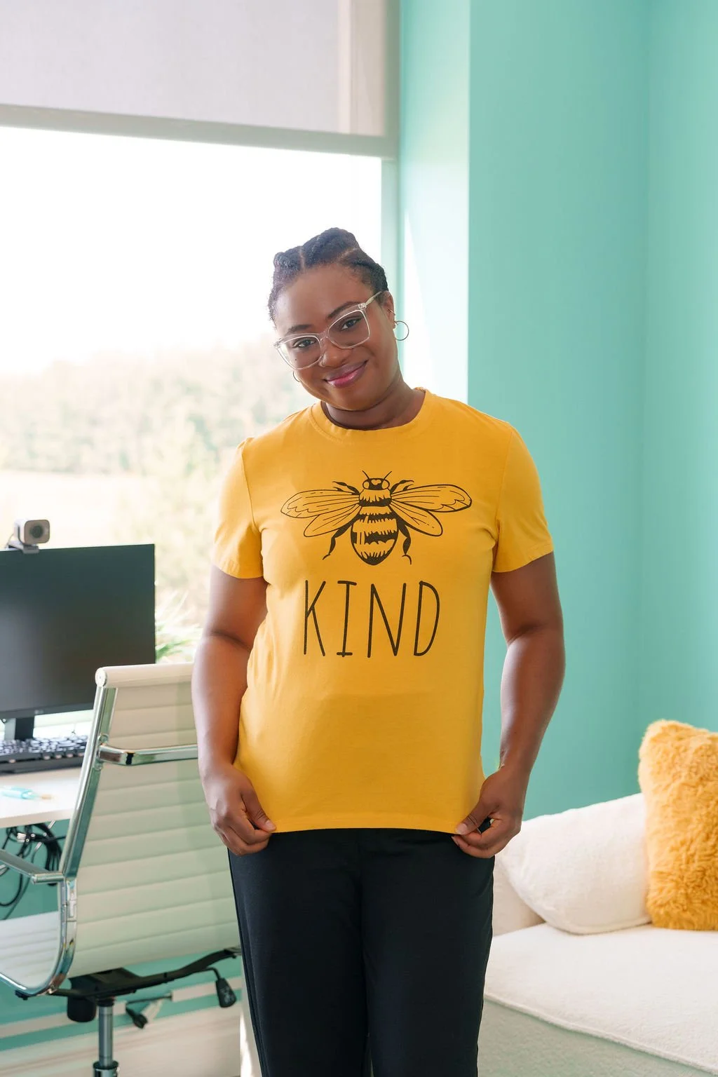 Woman smiling in an office, wearing a yellow t-shirt with a bee graphic and the word 'KIND', standing near a window with a computer and a white chair in the background.