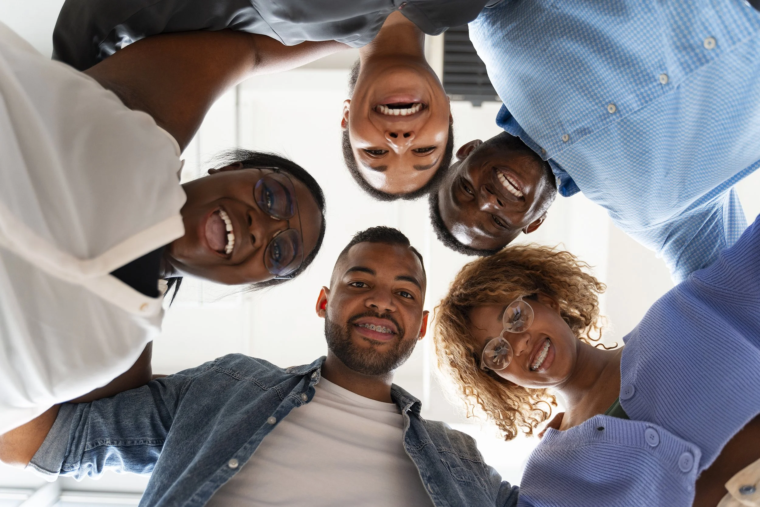 A group of six diverse people, smiling and laughing, forming a circle and looking down at the camera in a bright indoor setting.