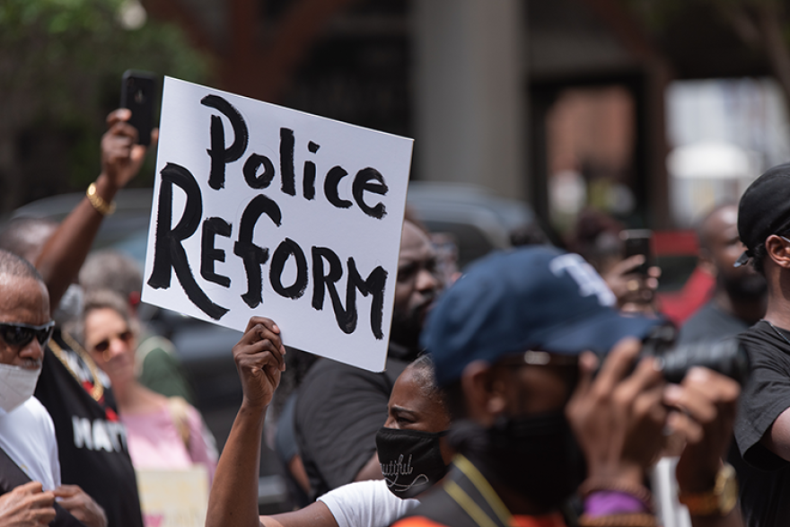 Protesters gathered outdoors, with one person holding a sign that reads 'Police Reform', amidst other demonstrators wearing masks and taking photos.