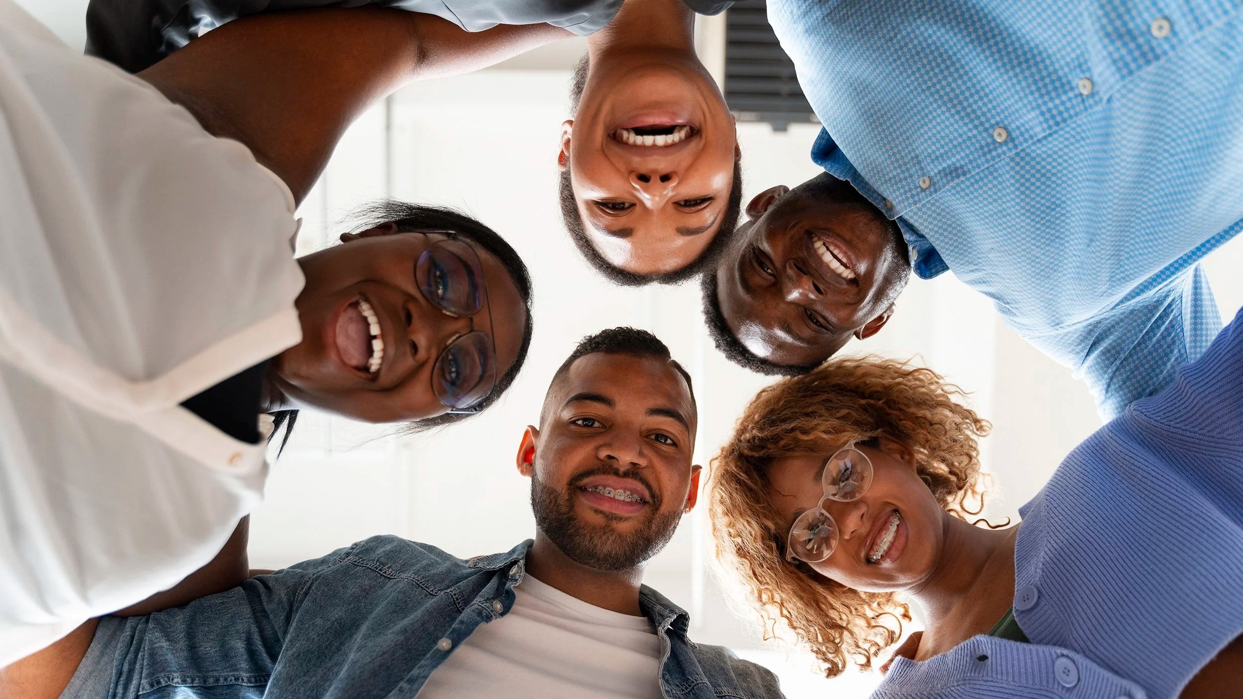 A diverse group of six happy people looking down into a camera, forming a circle with their heads tilted inward. They are smiling and seem to be enjoying a moment together.